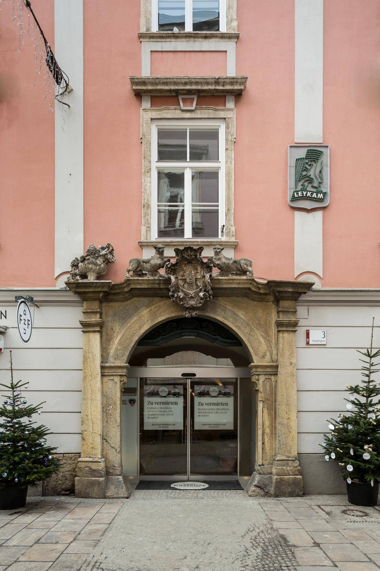 Historischer Eingangsbereich mit einem verzierten Steinbogen und großen Fenstern, flankiert von kleinen Weihnachtsbäumen.