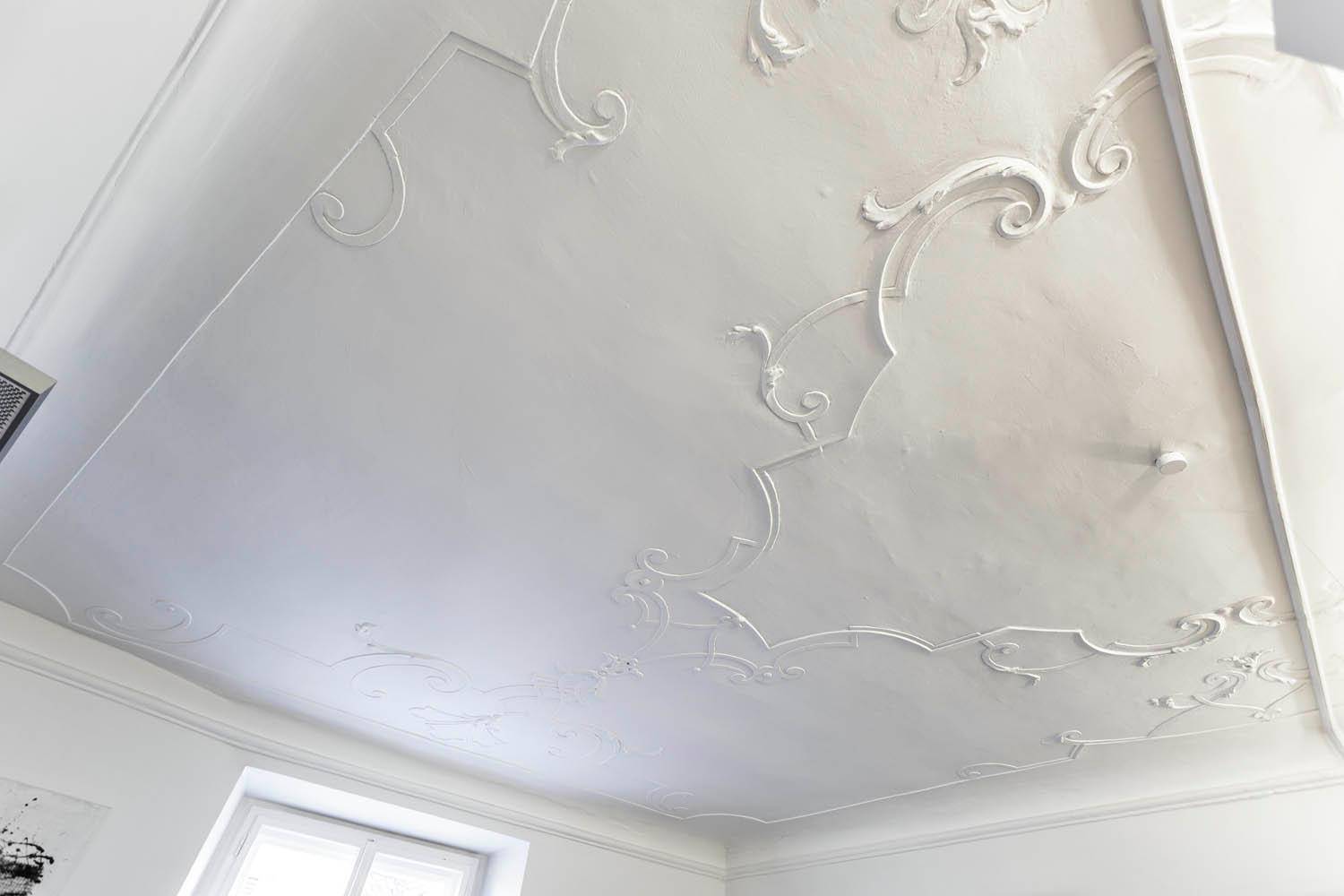 Weiße Decke mit detaillierten Stuckornamenten und einem Fenster, das natürliches Licht in den Raum bringt.