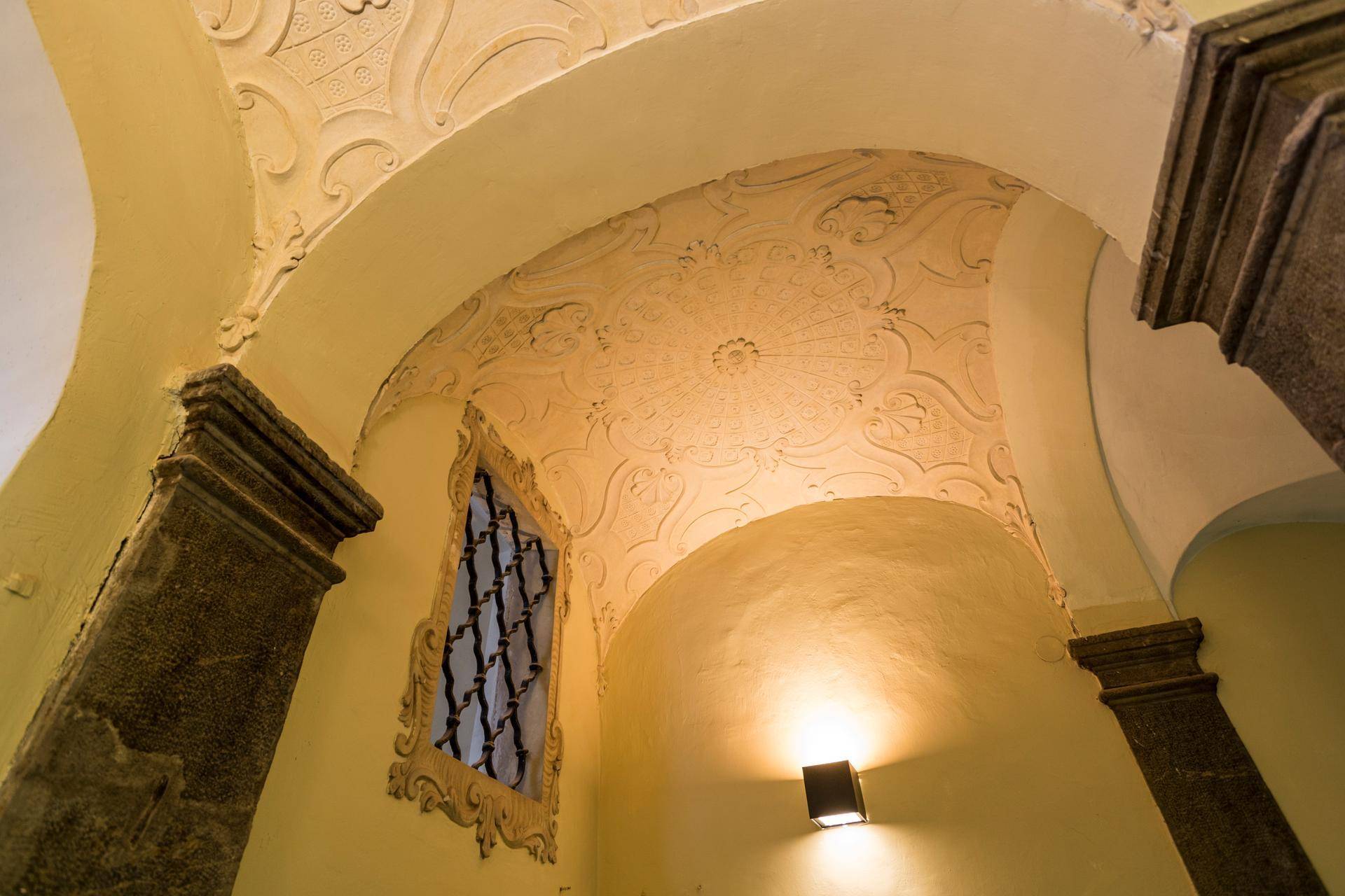 Historische Gewölbedecke mit aufwendigen Stuckverzierungen und einem kleinen Fenster, beleuchtet von einer Wandleuchte.