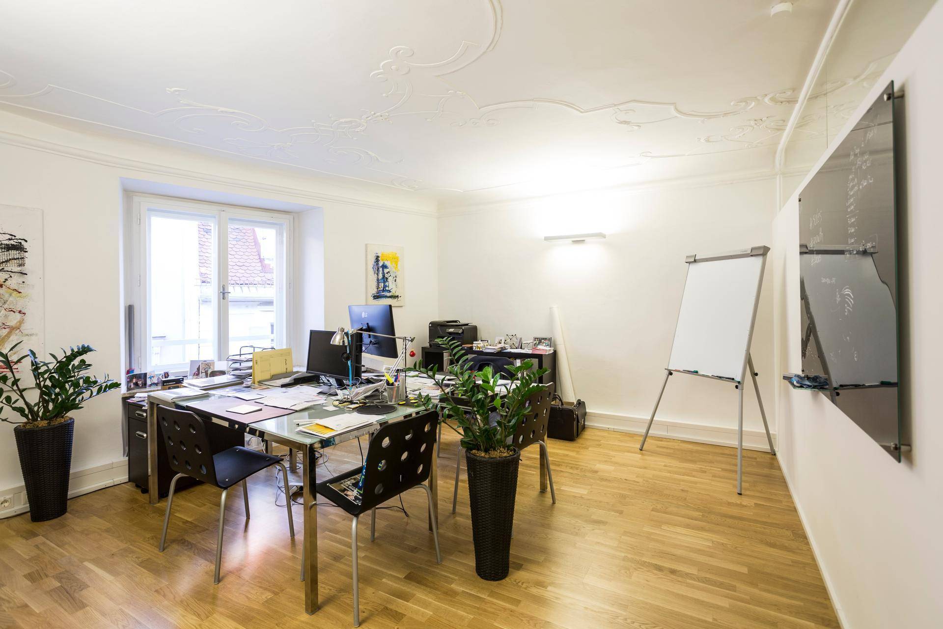 Helles Büro mit Stuckdecke, großem Fenster und Holzboden, eingerichtet mit Schreibtischen und Pflanzen.