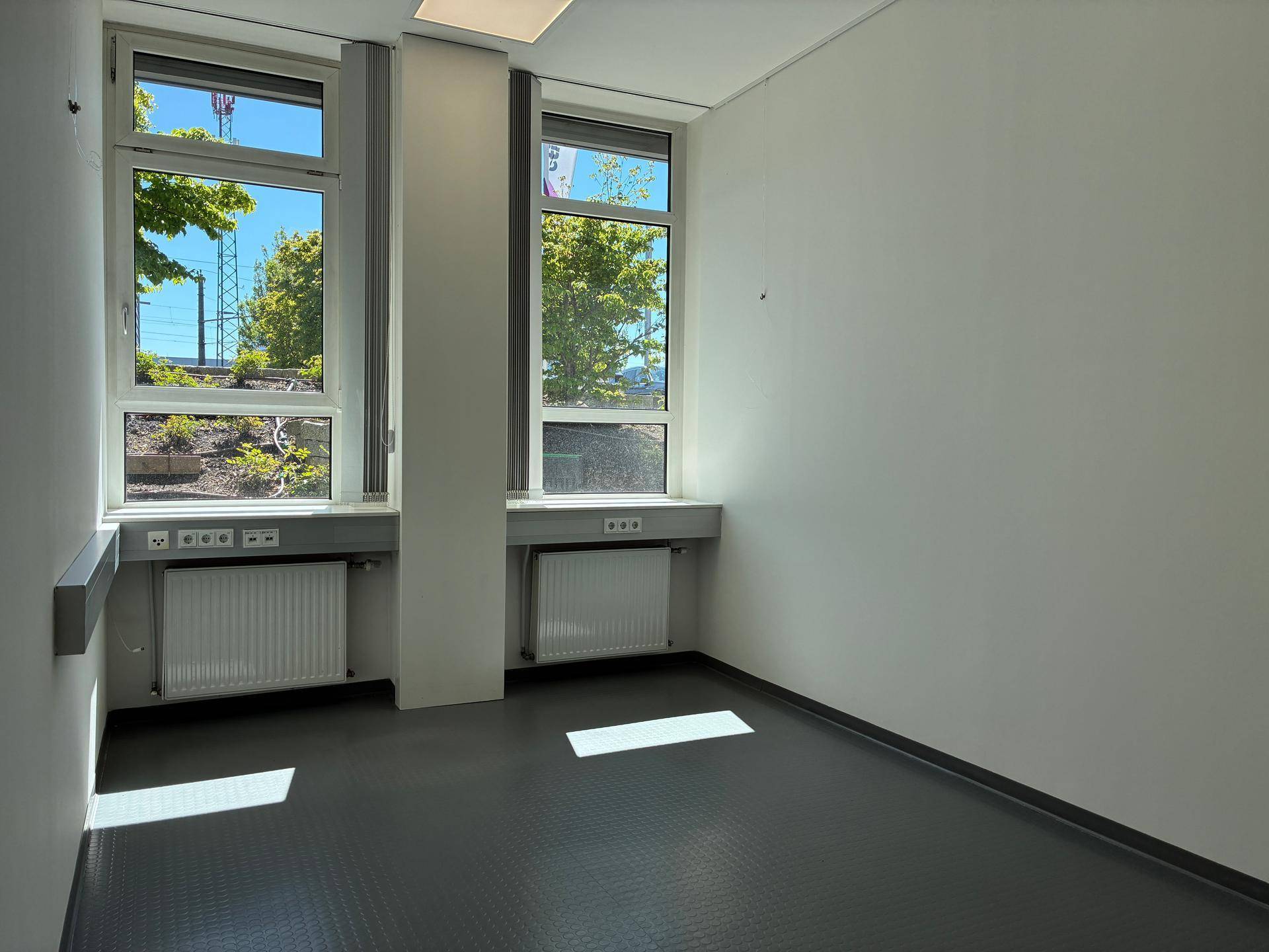 Heller Büroraum mit zwei großen Fenstern, die viel Tageslicht hereinlassen und einen Blick nach draußen bieten.