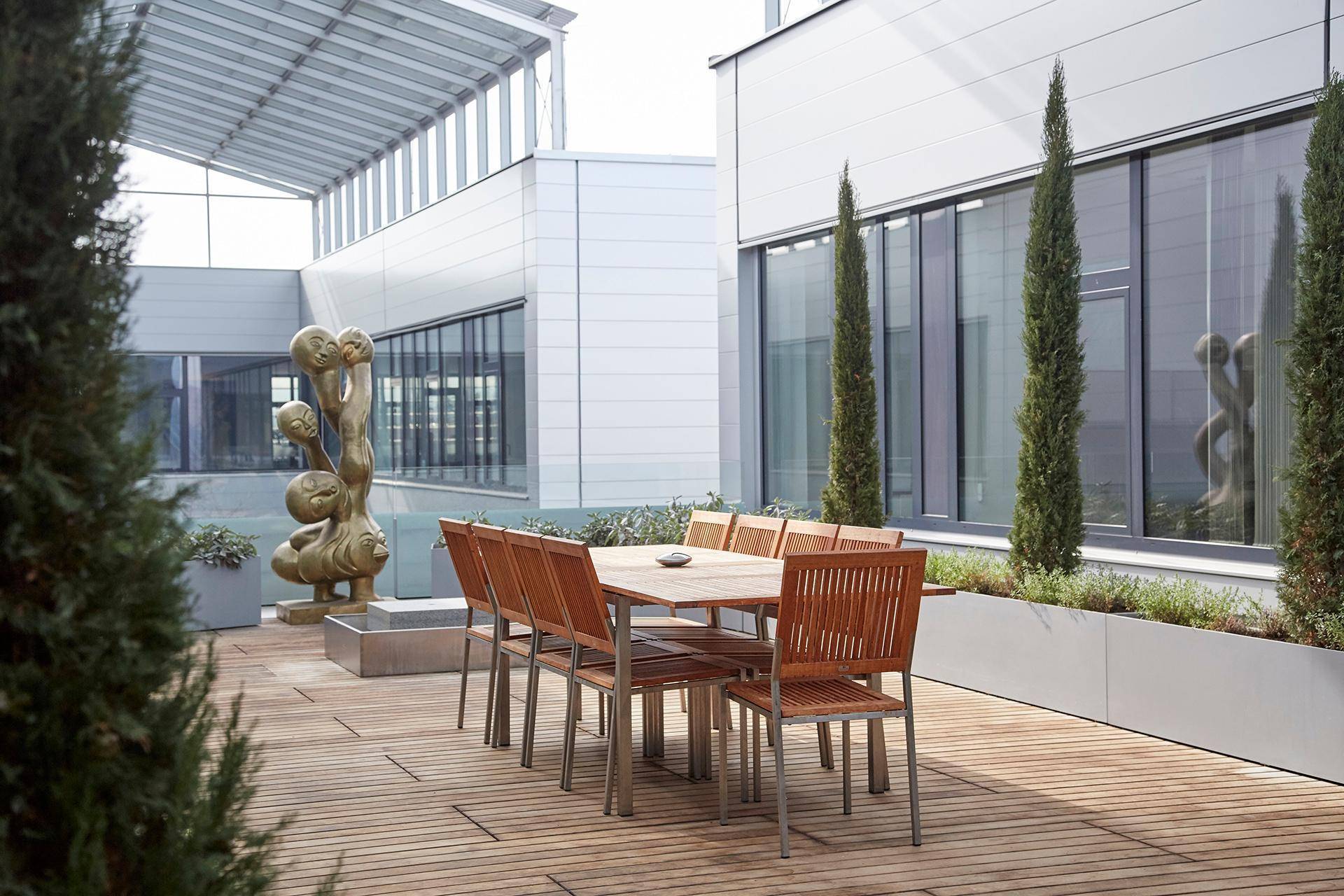 Geräumige Dachterrasse mit Holzdeck, Esstisch, hohen Bäumen in Pflanzgefäßen und einer modernen Skulptur.