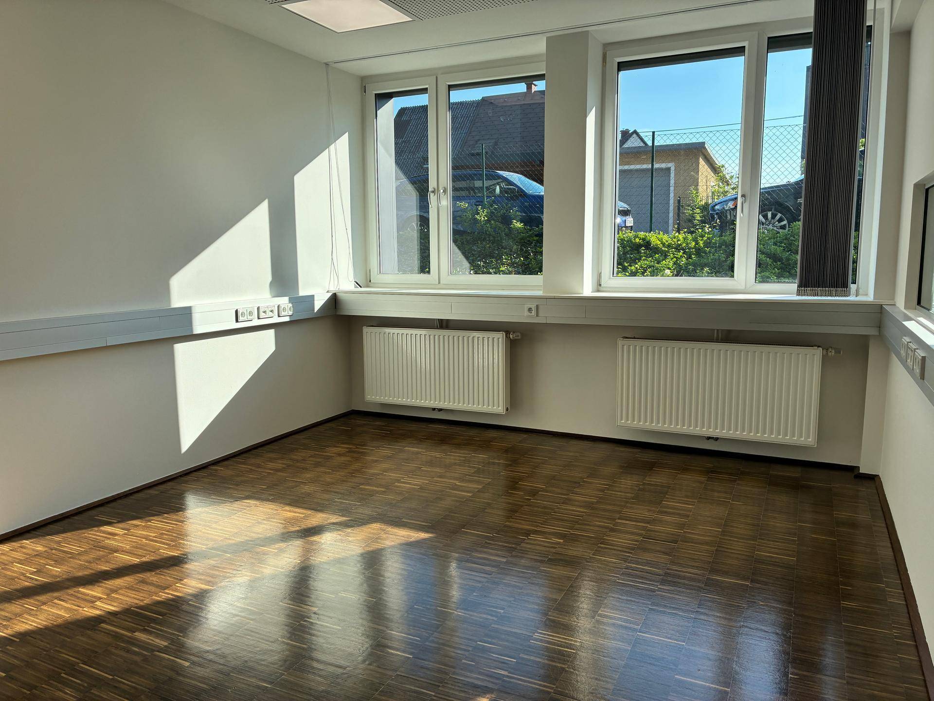 Leeres Büro mit großen Fenstern, Holzboden und Heizkörpern, das viel Tageslicht hereinlässt.