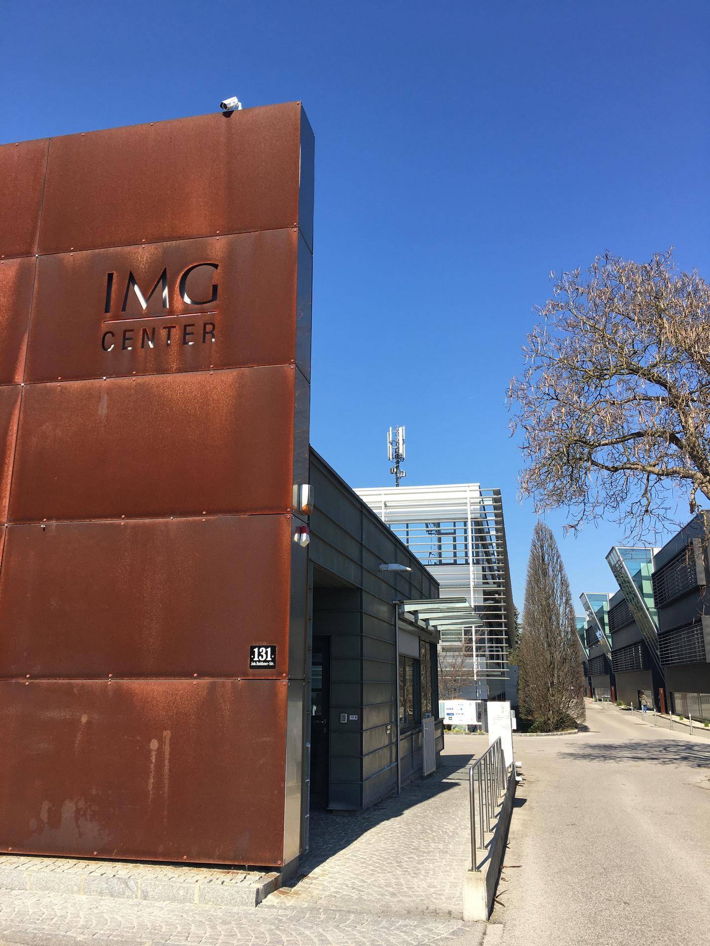 Eingangsbereich des IMG Centers mit markanter rostfarbener Fassade und moderner Architektur unter blauem Himmel.