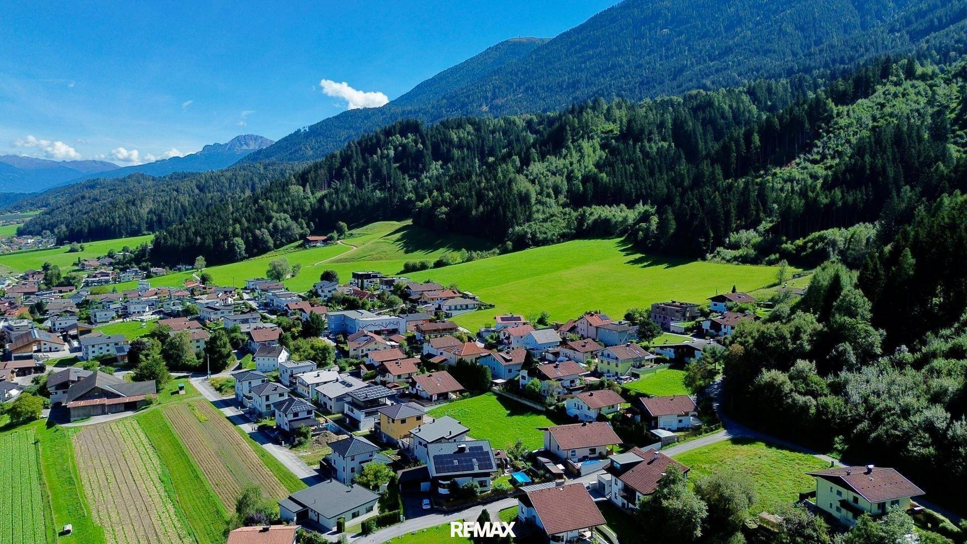 Weitläufige Luftaufnahme eines Dorfes in einem Tal, umgeben von grünen Feldern und bewaldeten Bergen.