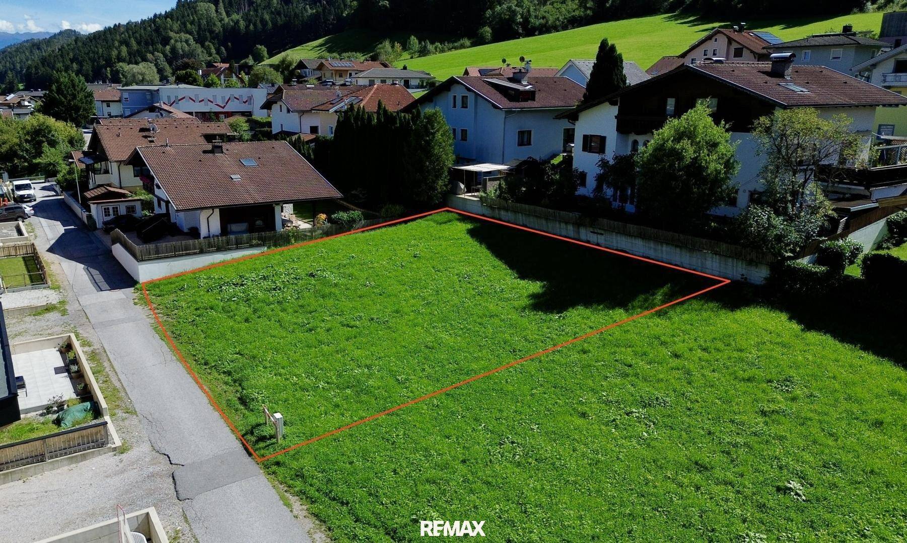 Nahaufnahme des grünen Baugrundstücks aus der Luft, umgeben von Häusern und mit Bergblick.