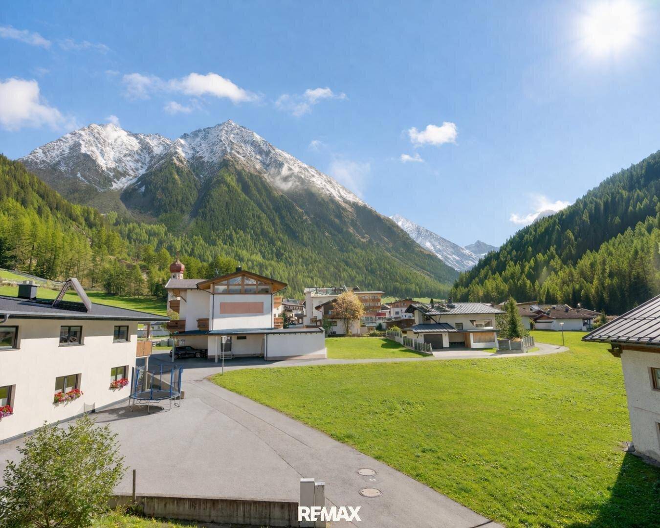 Malerisches Dorf in einem sonnigen Tal, umgeben von schneebedeckten Bergen und grünen Wiesen.