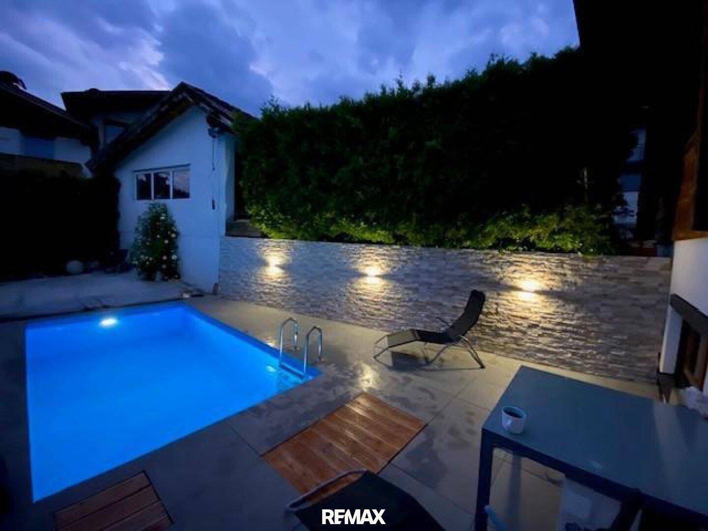 Beleuchteter Poolbereich bei Nacht, umgeben von einer Steinmauer und Hecke.