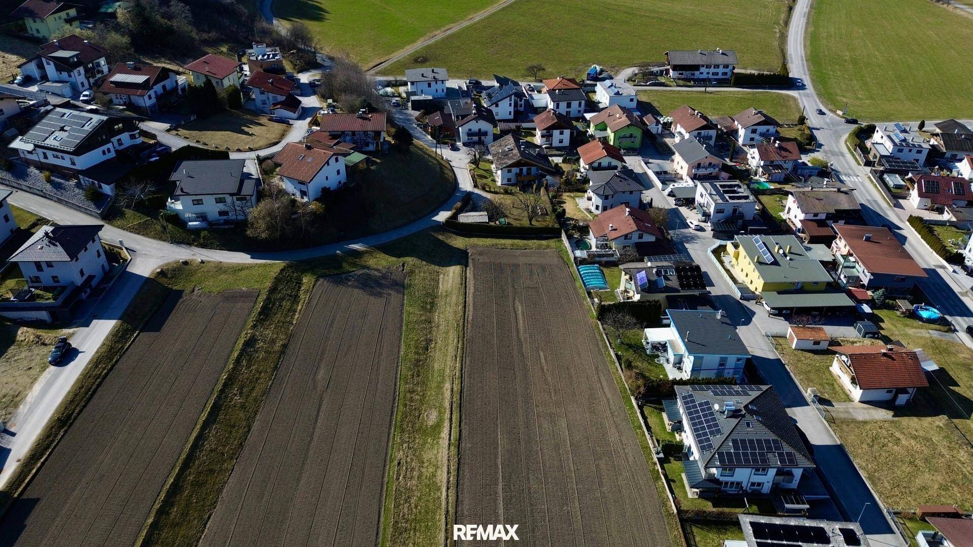 Luftaufnahme einer Wohnsiedlung mit angrenzenden Feldern und einer kurvigen Straße in sonniger Landschaft.