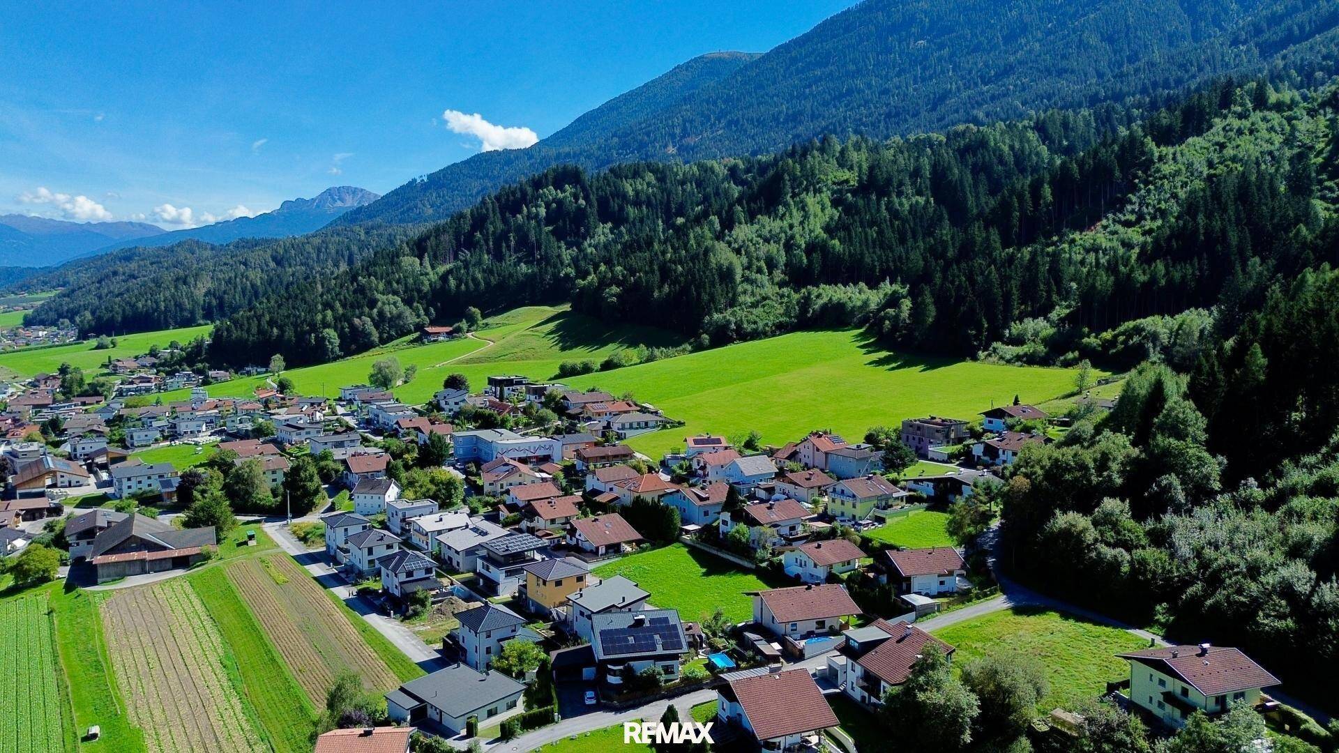 Weitläufige Ansicht eines Dorfes in einem grünen Tal, umgeben von bewaldeten Bergen unter blauem Himmel.