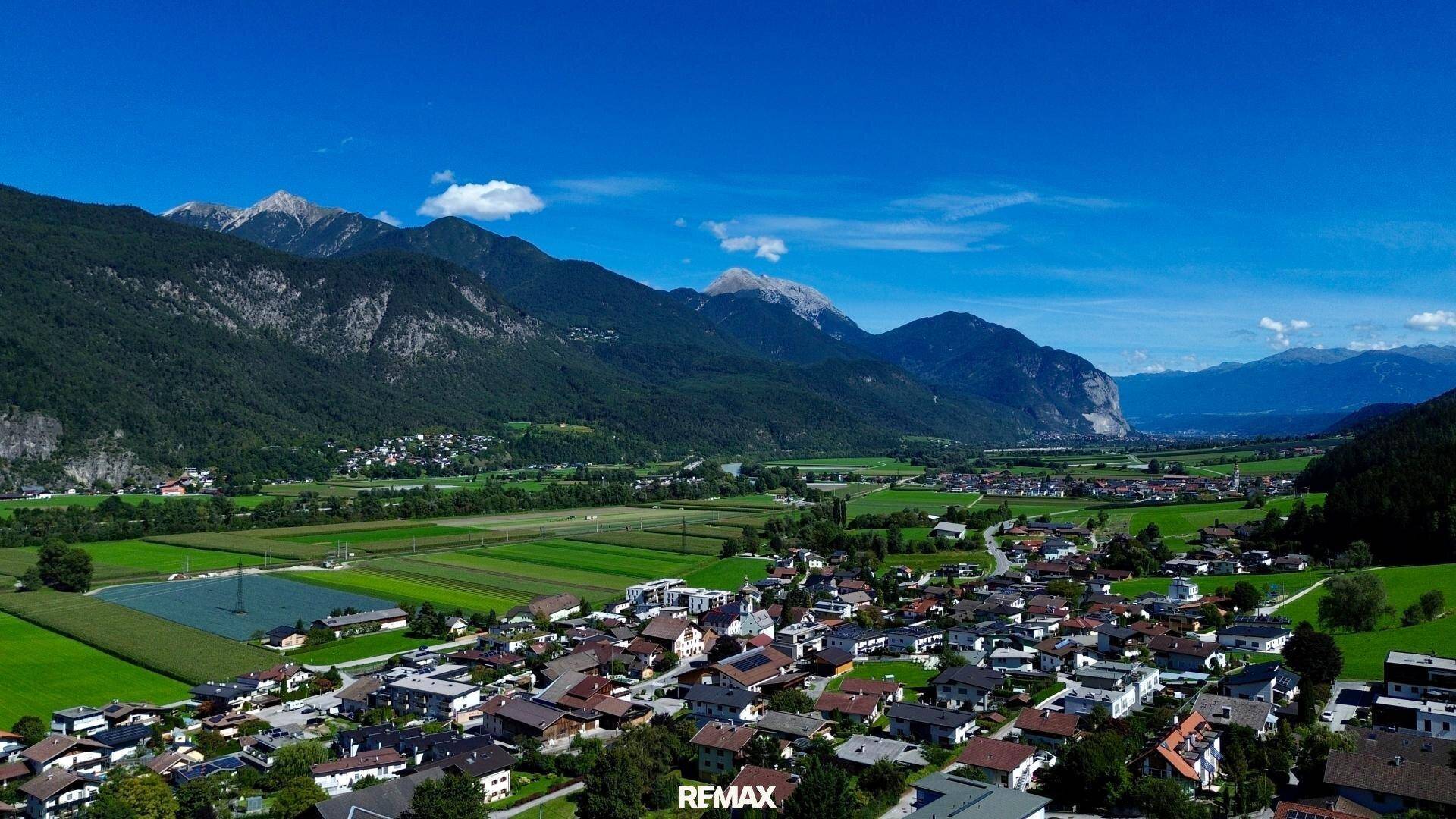 Panoramablick auf eine Siedlung im Tal, umrahmt von majestätischen Bergen und weiten Feldern.