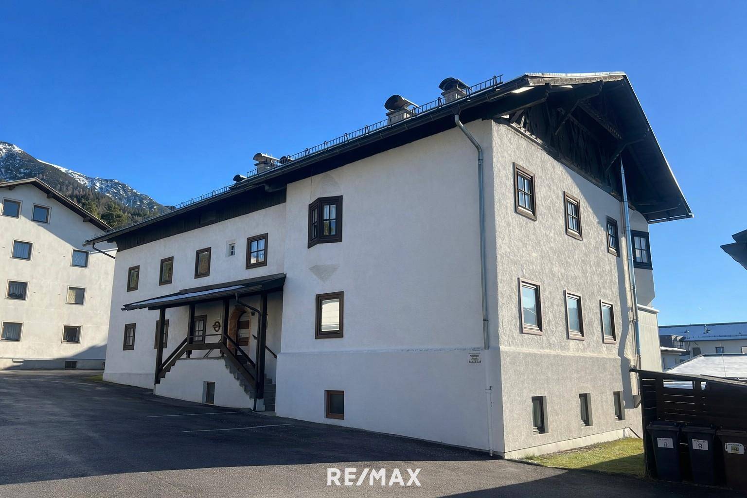 Traditionelles Gebäude mit weißer Fassade, dunklen Fenstern und alpiner Architektur vor Bergkulisse.