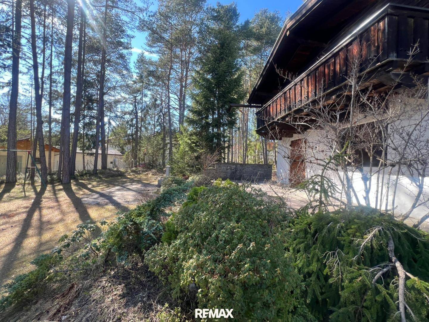 Seitenansicht eines Hauses mit Holzbalkon, umgeben von üppigem Grün und hohen Bäumen.