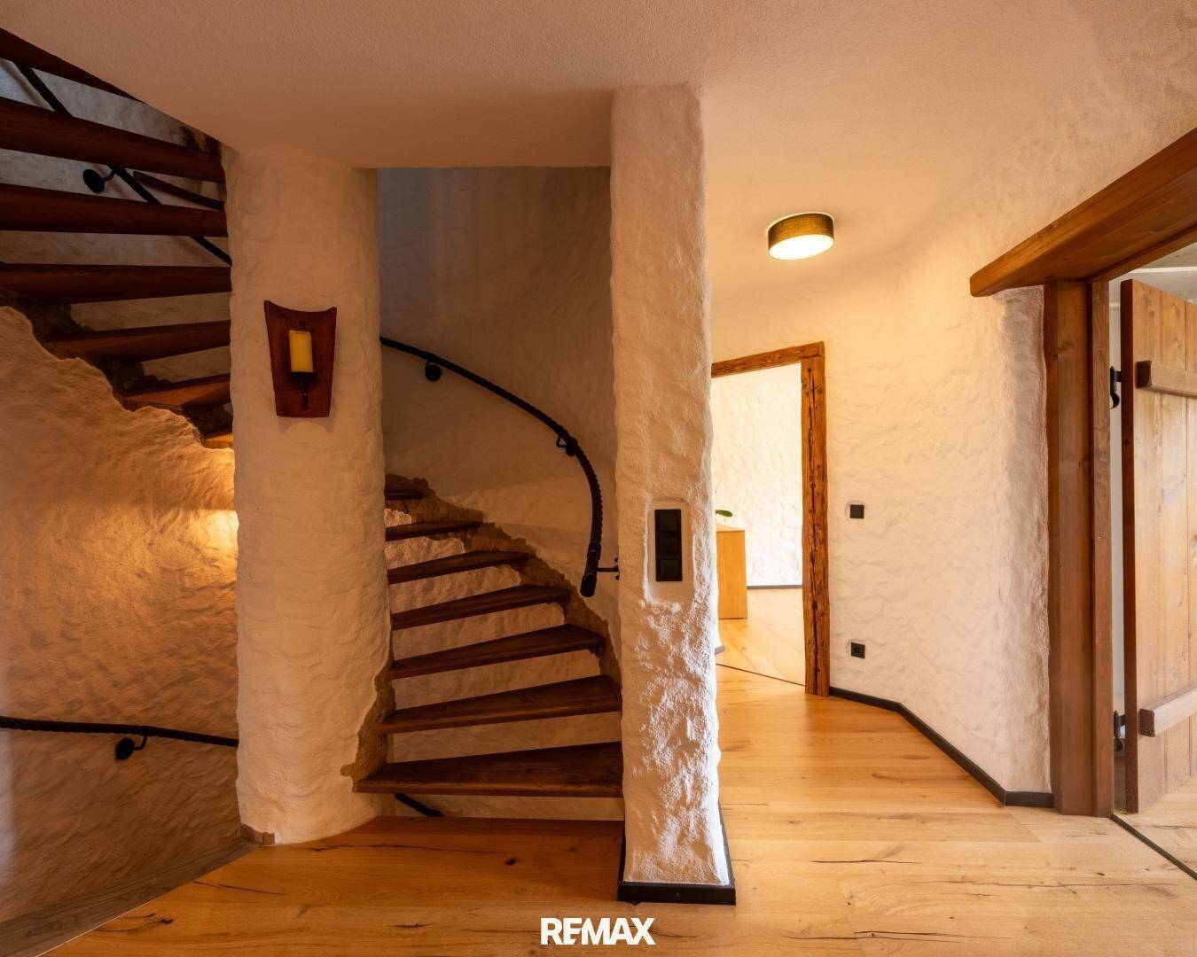 Heller Flur mit geschwungener Holztreppe und rustikalen Wänden, die zu weiteren Räumen führen.