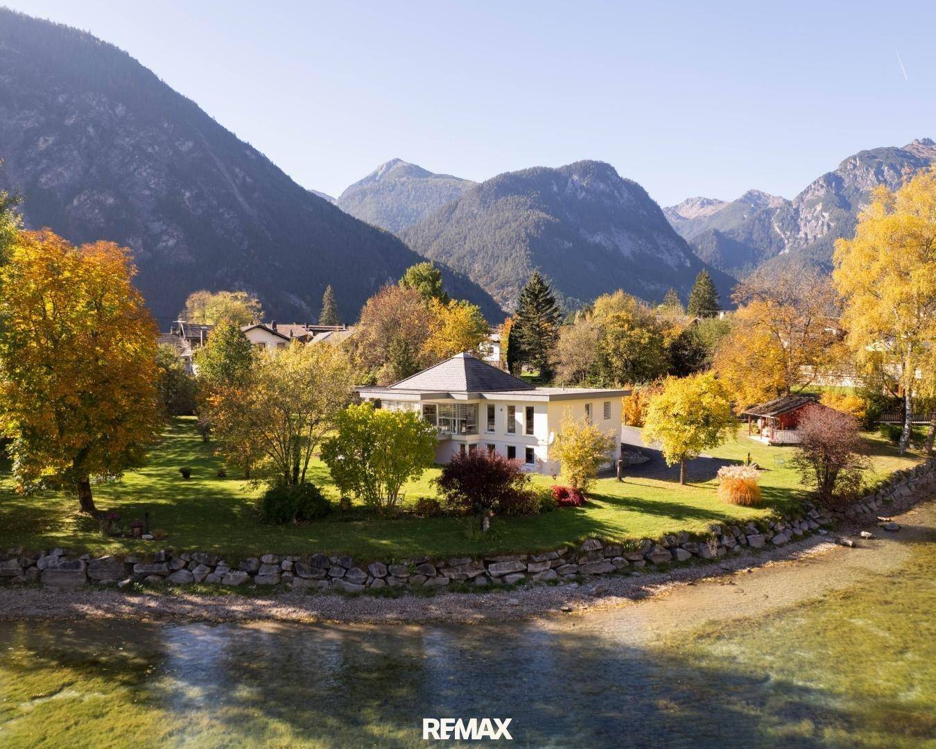 Moderne Villa am Ufer eines klaren Sees, umgeben von herbstlicher Vegetation und Bergen.