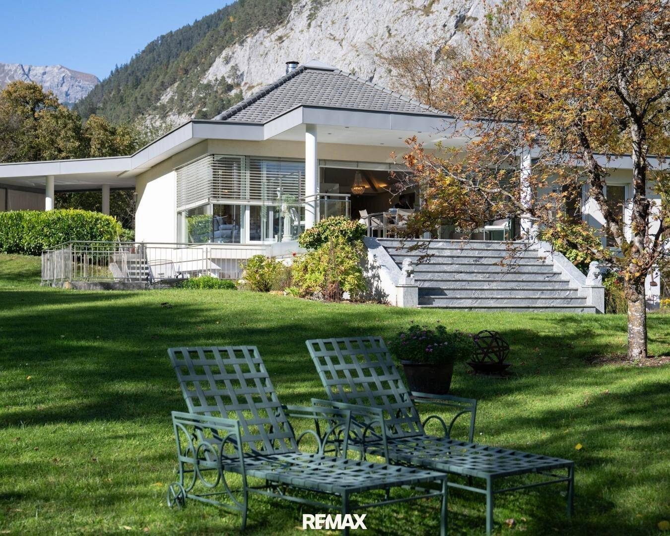 Entspannungsbereich im Garten mit Liegestühlen und Blick auf die moderne Villa und die Berge.