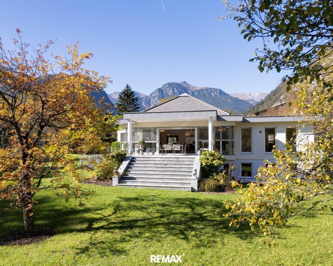 Elegantes Haus mit großer Terrasse und Treppe, umgeben von einem gepflegten Garten und Bergen.