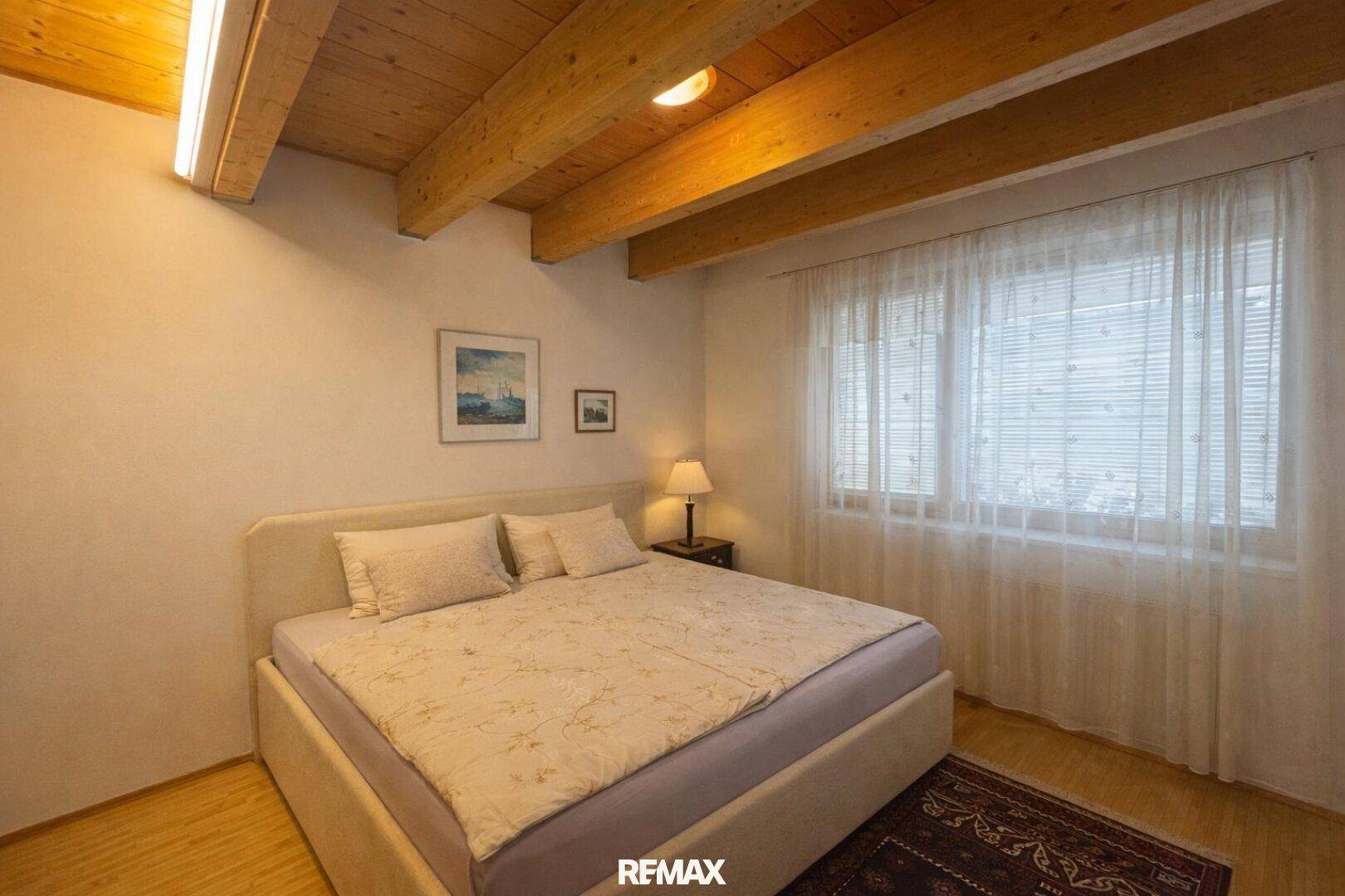 Helles Schlafzimmer mit Doppelbett, Holzbalkendecke und Fenster mit Vorhängen.