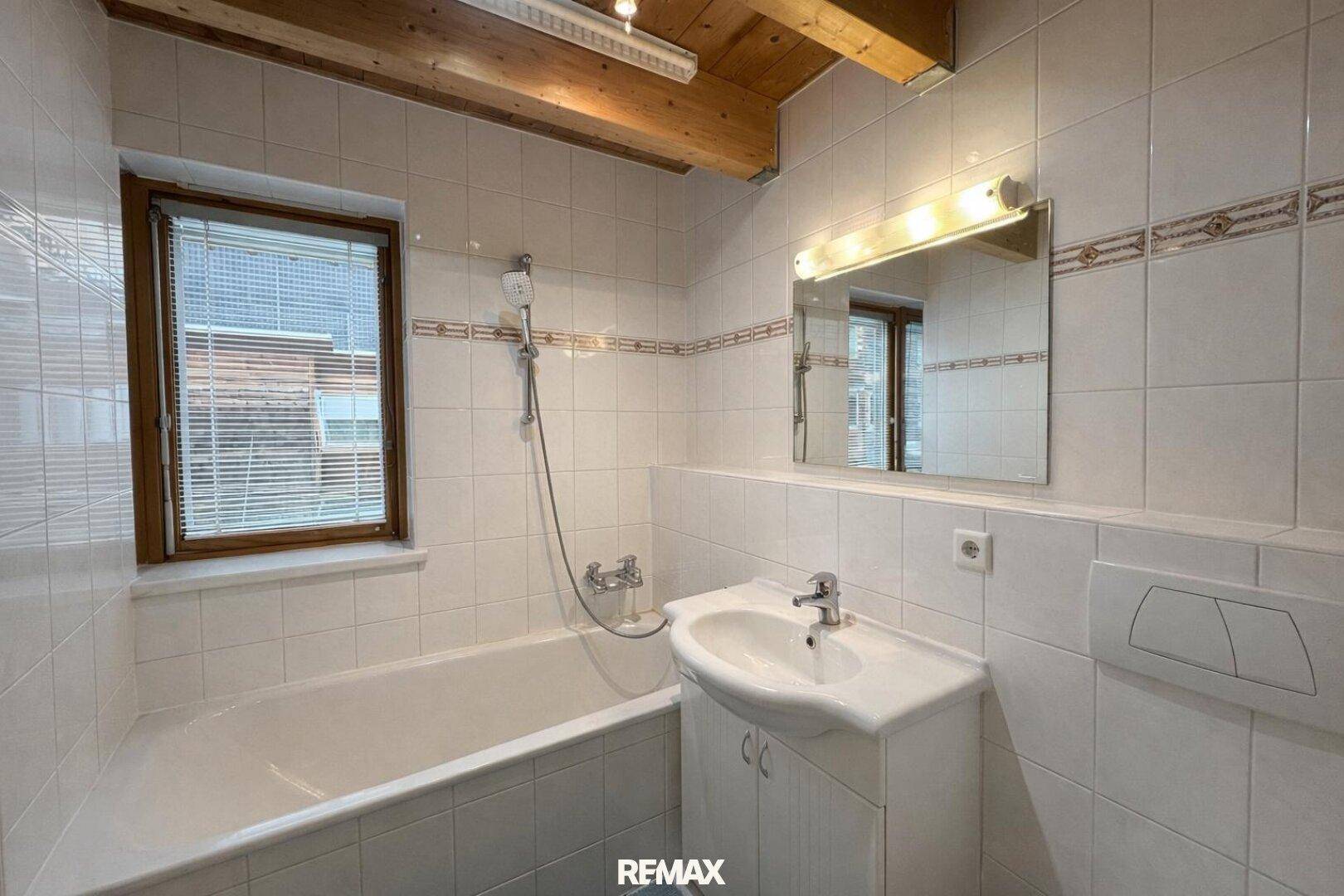 Gefliestes Badezimmer mit Badewanne, Waschtisch, Spiegel und Fenster unter Holzbalkendecke.