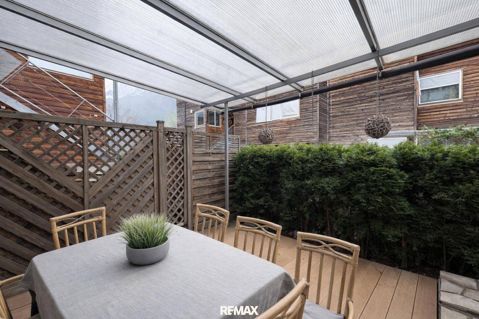 Überdachte Terrasse mit Esstisch, Holzdeck und grüner Hecke, ideal für Mahlzeiten im Freien.