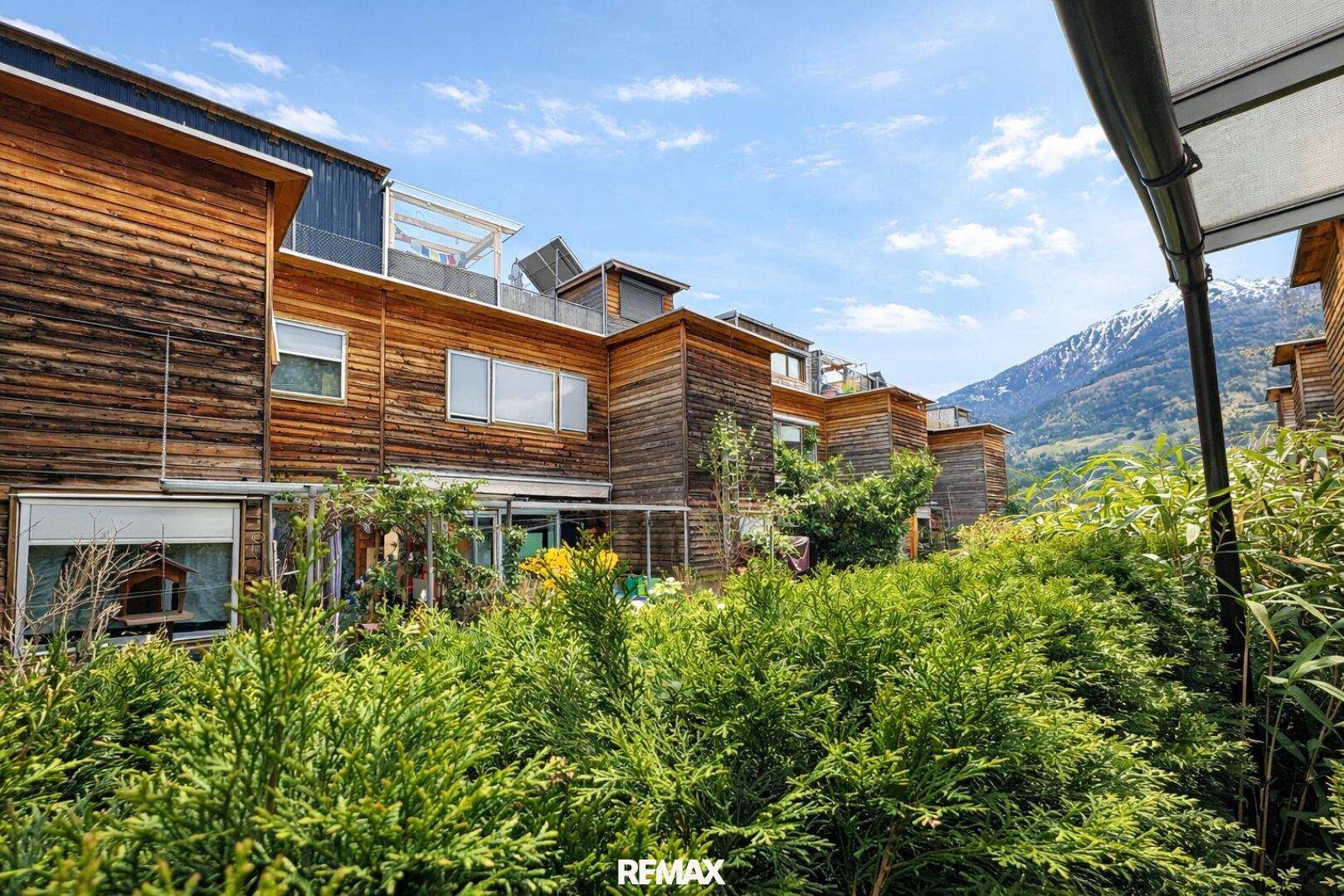 Üppiger Gartenbereich mit grünen Büschen, Holzfassade und Bergblick im Hintergrund.