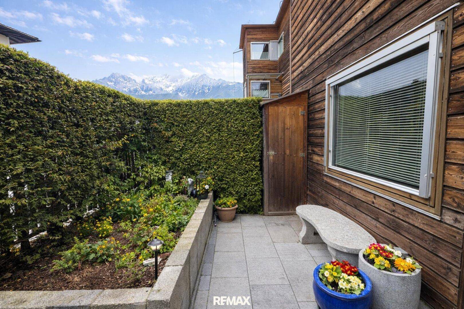 Kleiner, gepflasterter Gartenbereich mit Steinbank, hoher Hecke und Bergblick.