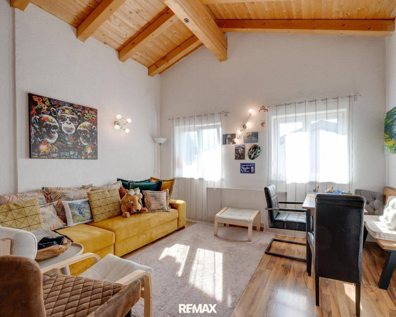 Helles Wohnzimmer mit sichtbaren Holzbalken, gelbem Sofa und Teppichboden.