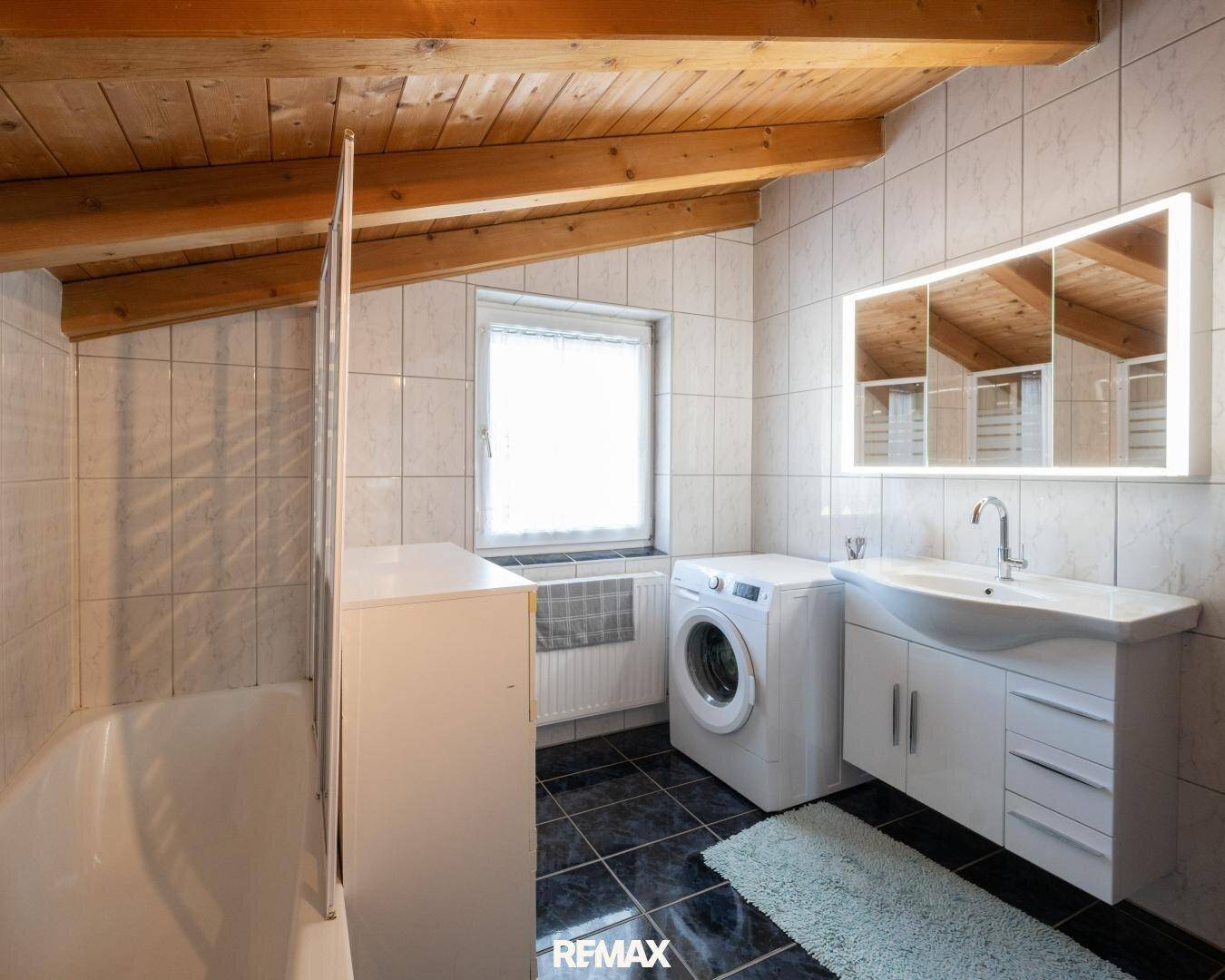 Badezimmer mit Holzbalkendecke, Waschmaschine, Waschtisch und Duschkabine.