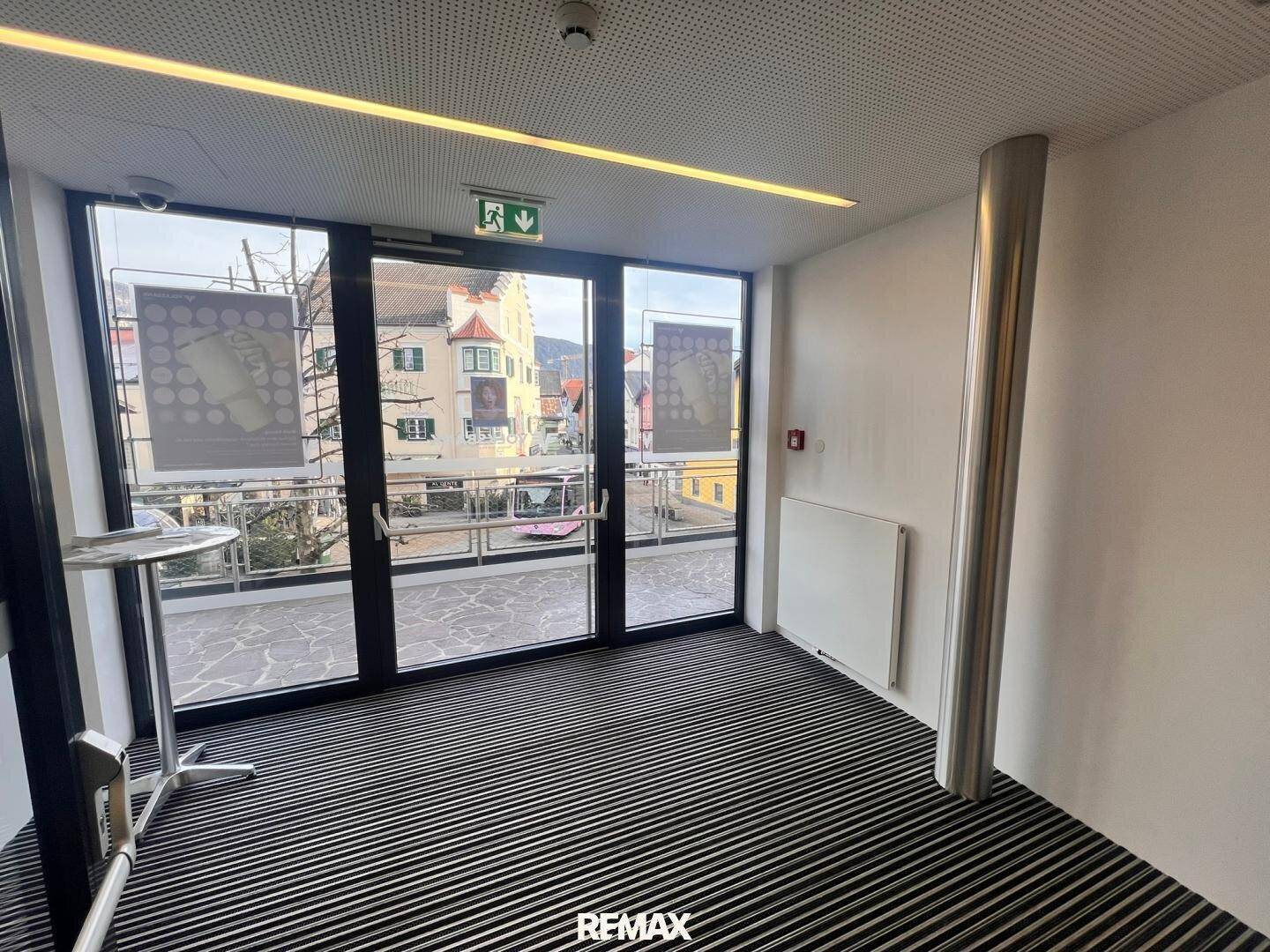 Heller Büroraum mit großen Fenstern, die auf eine Terrasse blicken, und gestreiftem Teppichboden.