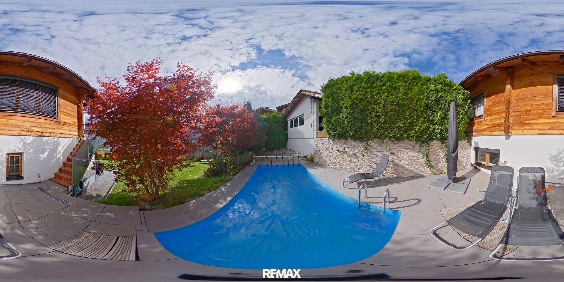 Außenbereich mit Pool, Liegestühlen und umgebender Bepflanzung unter blauem Himmel.
