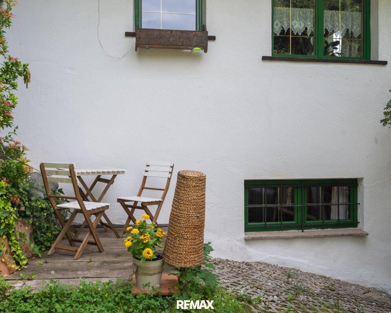 Kleiner Außenbereich mit Tisch und Stühlen vor einer weißen Hauswand mit grünen Fenstern.
