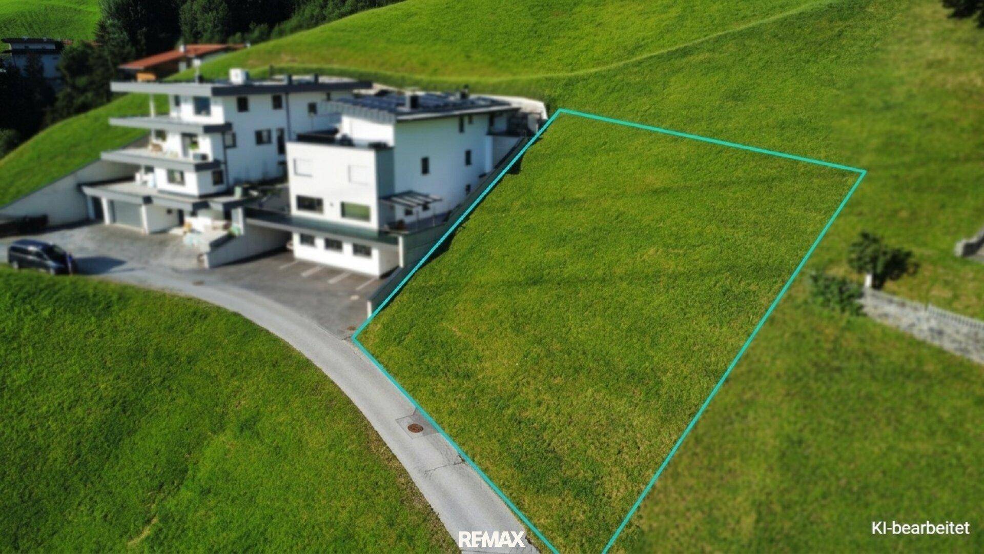 Luftaufnahme eines sonnigen Baugrundstücks mit blauer Umrandung neben einem modernen Gebäude am Hang.