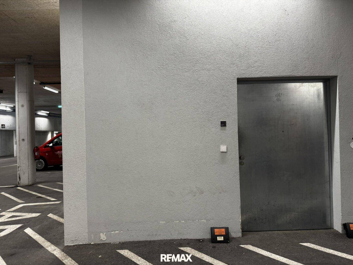 Tiefgarage mit grauen Wänden, einer Stahltür und einem geparkten roten Auto im Hintergrund.