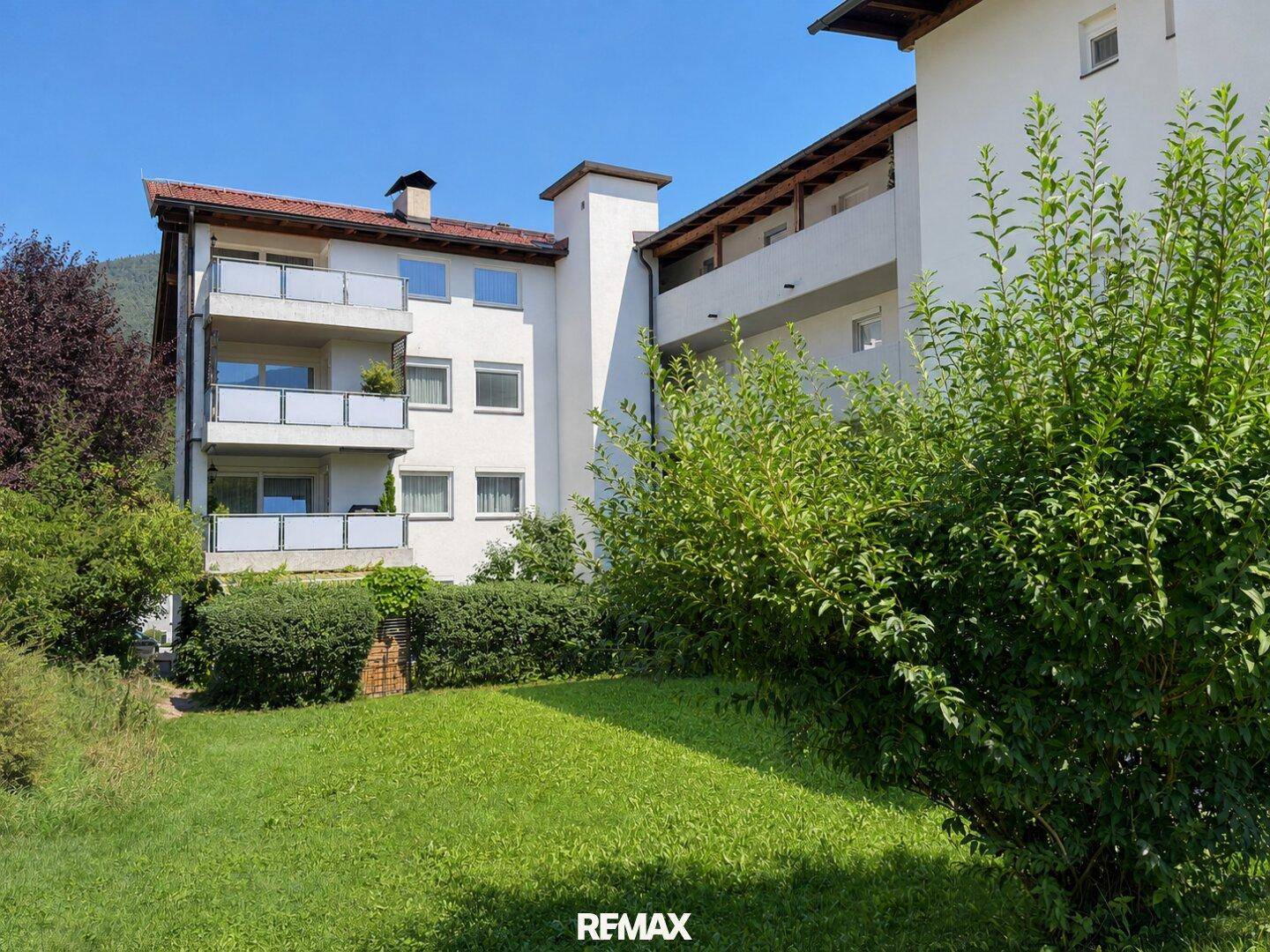 Mehrfamilienhaus mit mehreren Balkonen, umgeben von viel Grün und mit Bergblick in der Ferne.