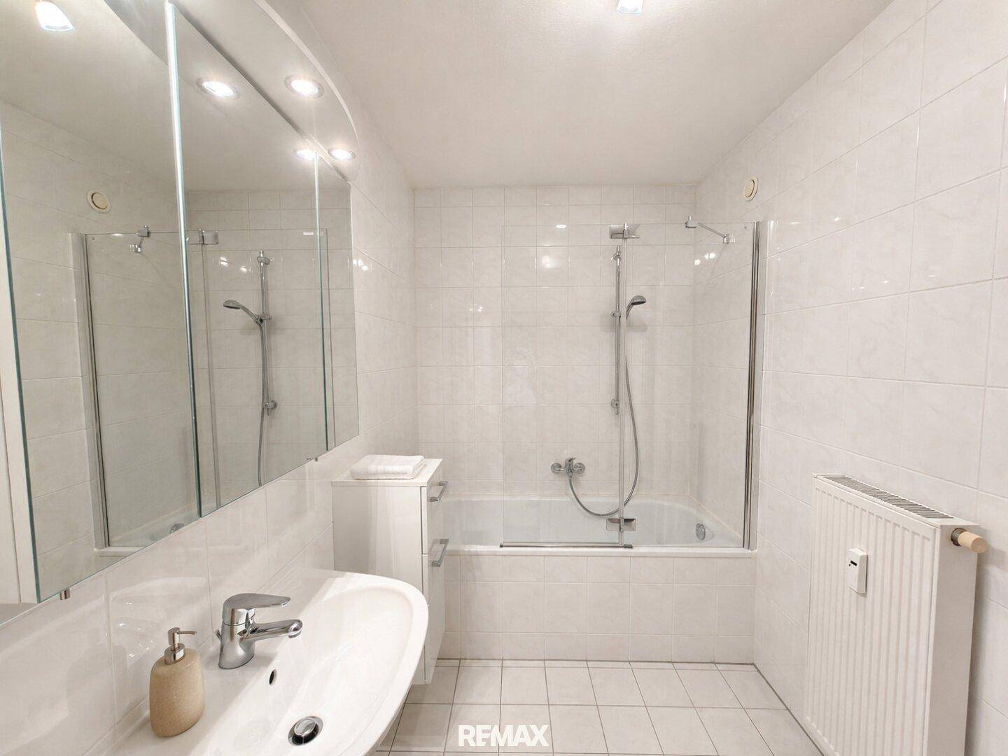 Helles Badezimmer mit weißem Fliesenboden, Waschbecken, großem Spiegelschrank und Badewanne mit Duschfunktion.
