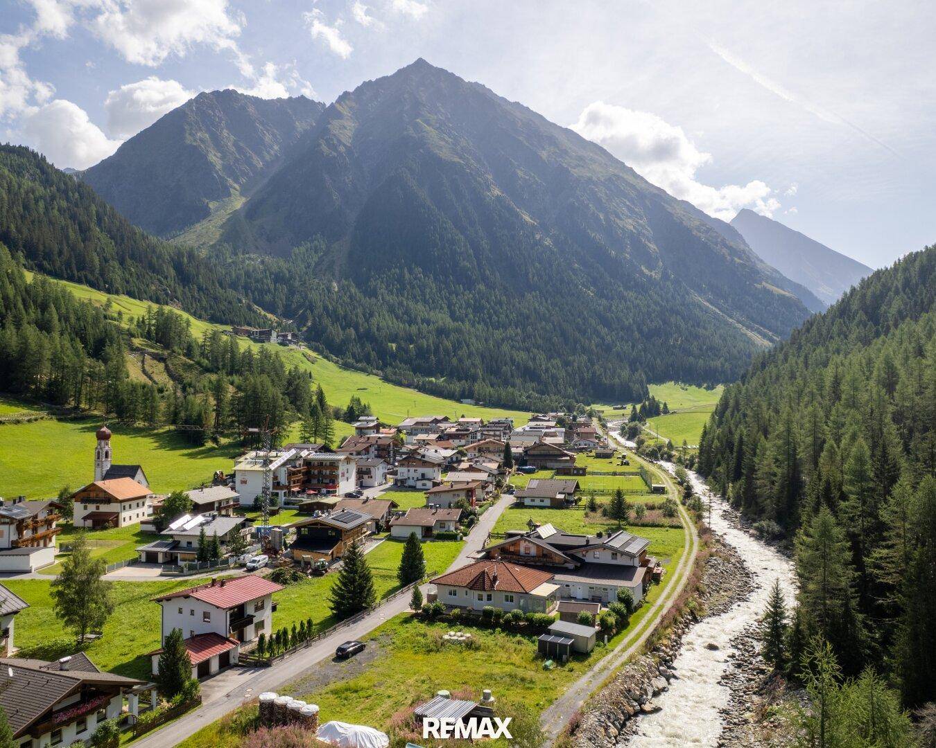 Luftaufnahme eines malerischen Dorfes in einem Tal, umgeben von Bergen und einem fließenden Fluss.