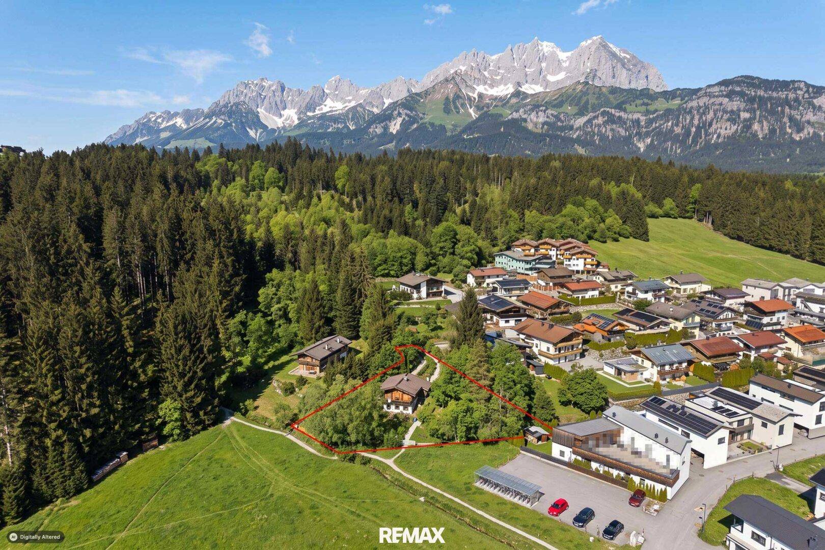 Weitläufige Luftaufnahme des traditionellen Chalets mit Grundstück und angrenzendem Dorf vor Bergpanorama.