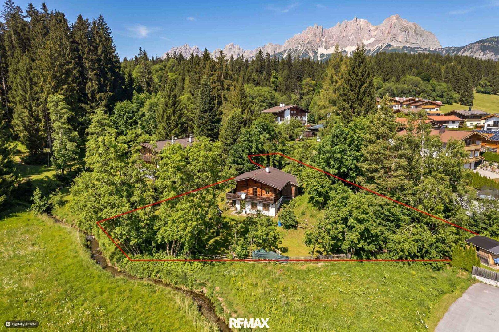 Traditionelles Holzchalet mit Grundstücksgrenzen markiert, umgeben von Wald und Wiesen mit Bergblick.