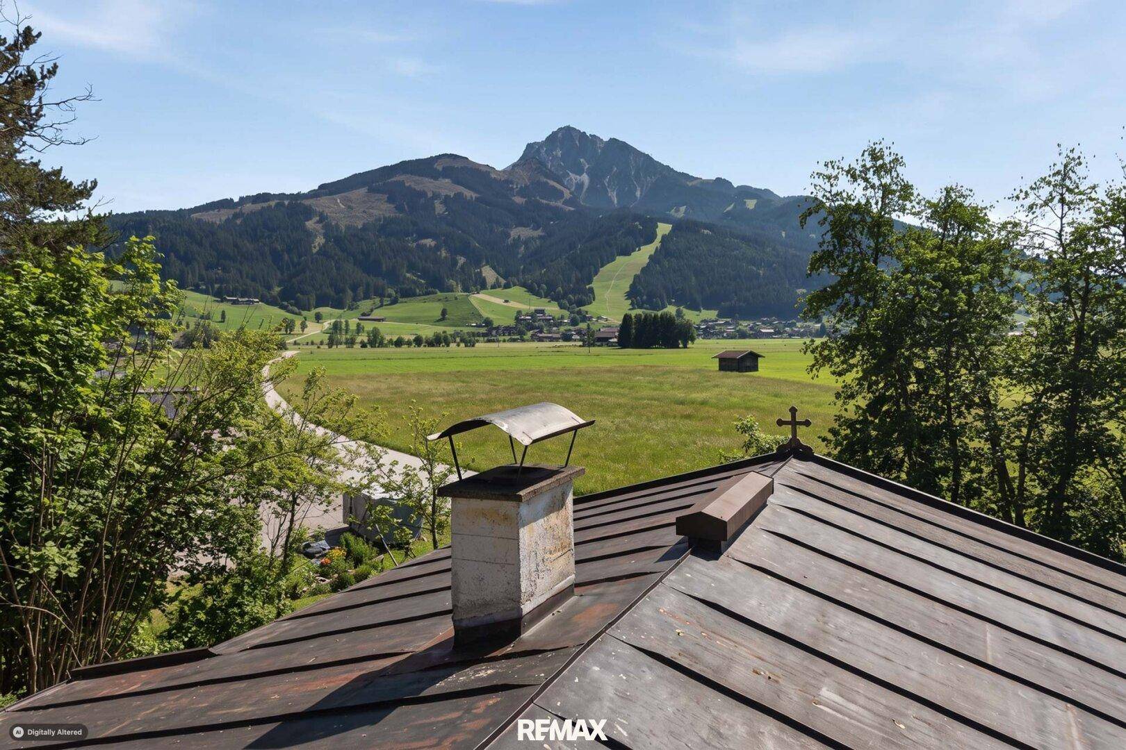 Blick über ein traditionelles Holzdach auf weite grüne Felder und eine beeindruckende Berglandschaft.