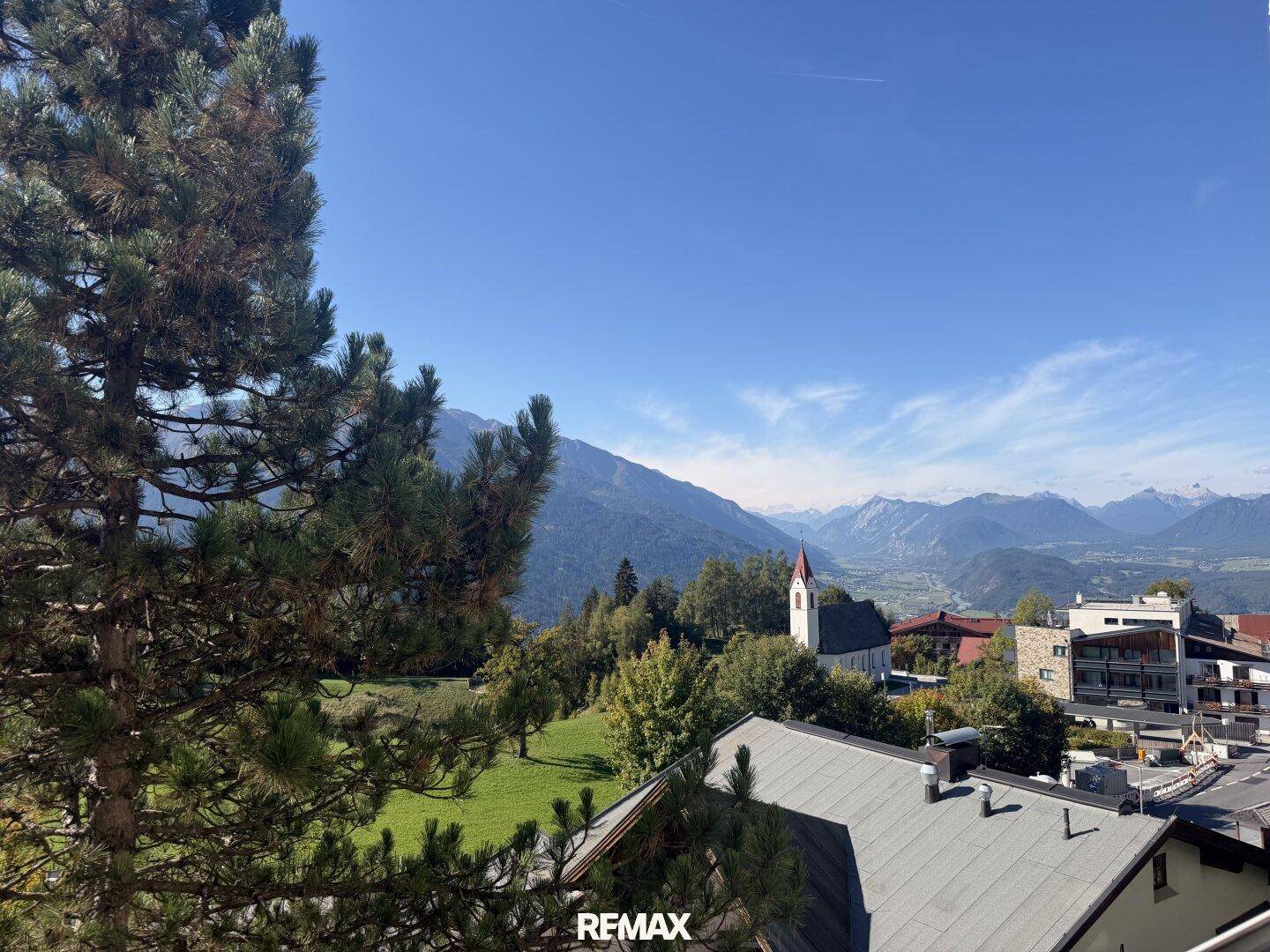 Panoramablick auf die umliegende Berglandschaft mit einer Kirche und grünen Hügeln unter blauem Himmel.