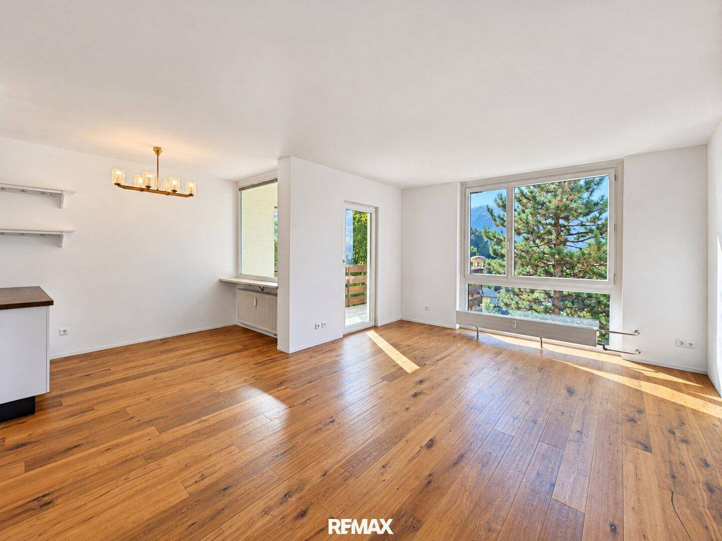 Geräumiger, leerer Wohnbereich mit Holzboden, großen Fenstern und Zugang zum Balkon mit Bergblick.