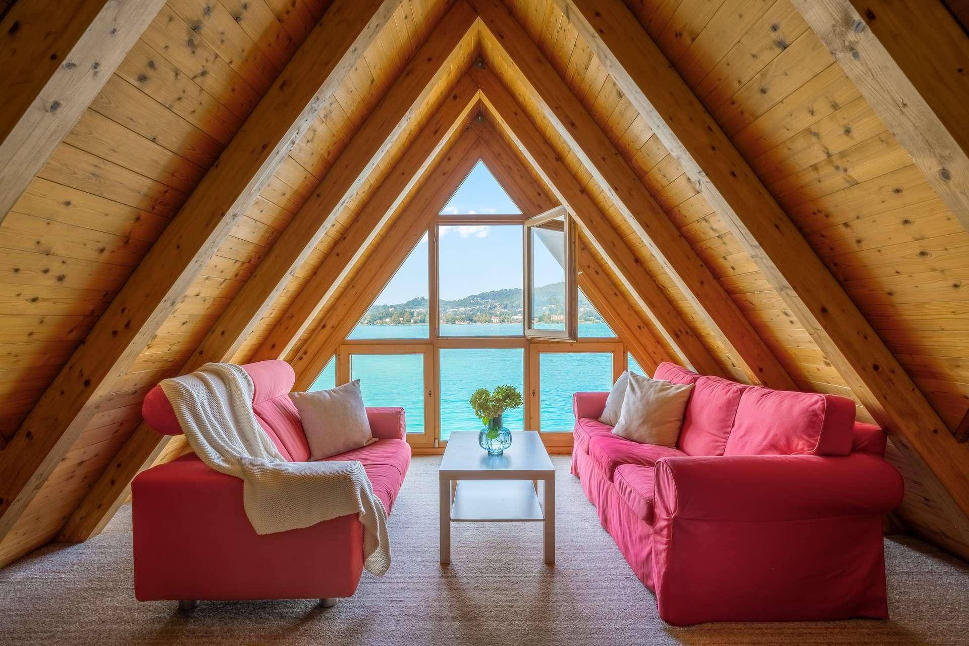 Gemütliches Wohnzimmer im Dachgeschoss mit hohen Holzdecken, zwei roten Sofas und Panoramablick auf den See.
