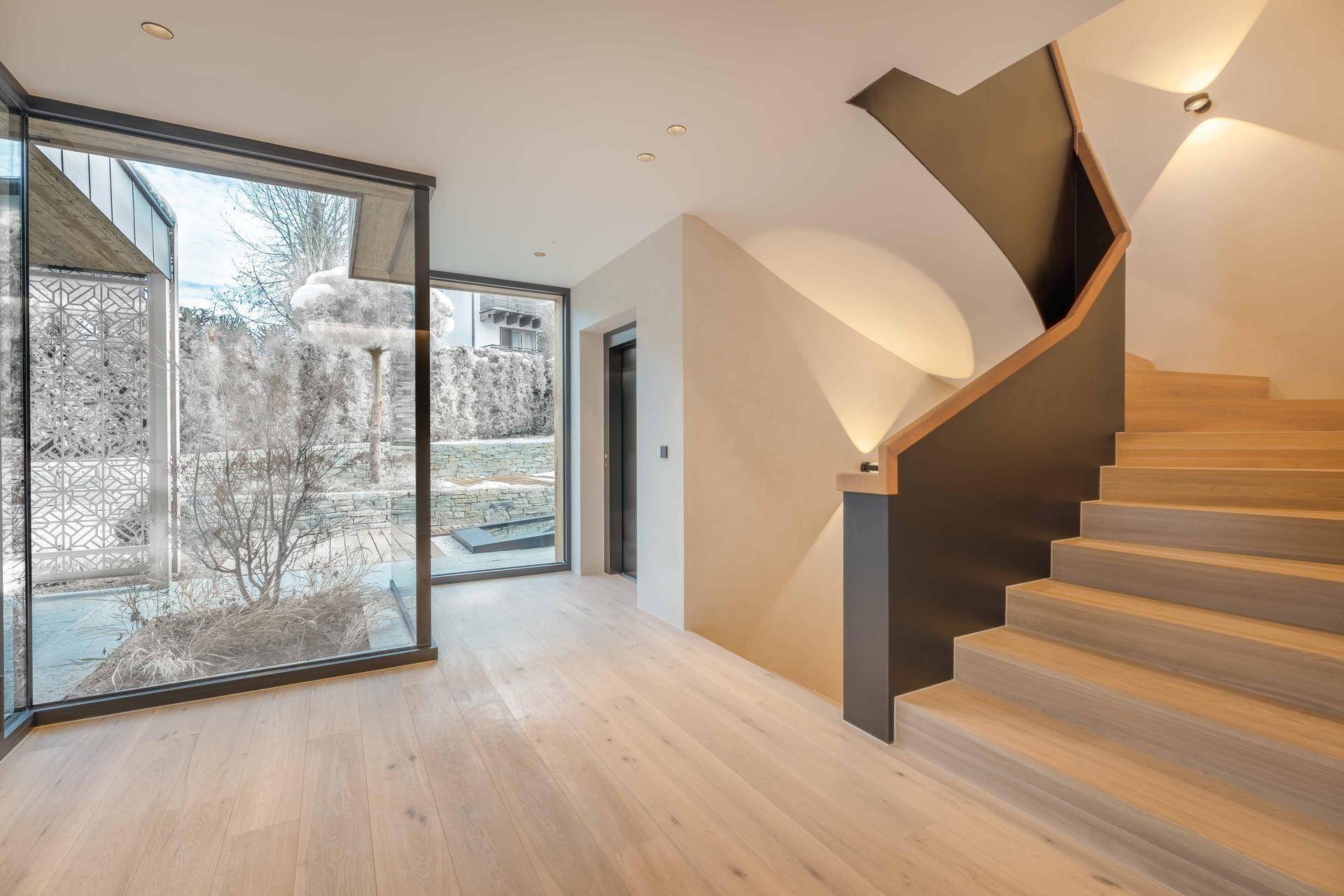 Heller Eingangsbereich mit moderner Holztreppe, großen Fenstern und Zugang zum Aufzug.