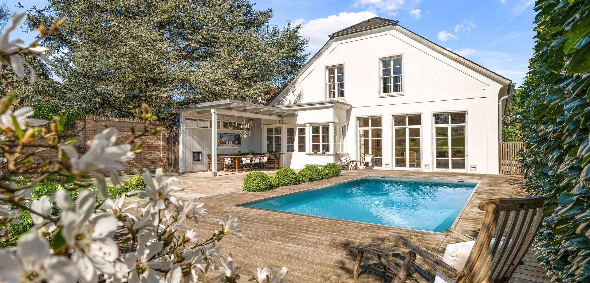 Rückseite der Villa mit großem Pool, Holzdeck und überdachtem Essbereich im gepflegten Garten.