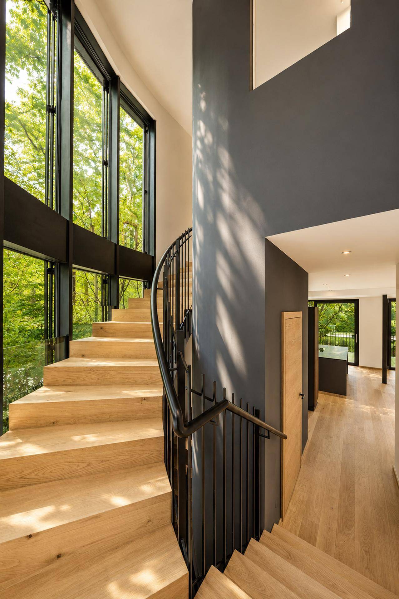 Elegantes Treppenhaus mit Holzstufen, geschwungenem Geländer und hohen Fenstern mit Blick auf Bäume.