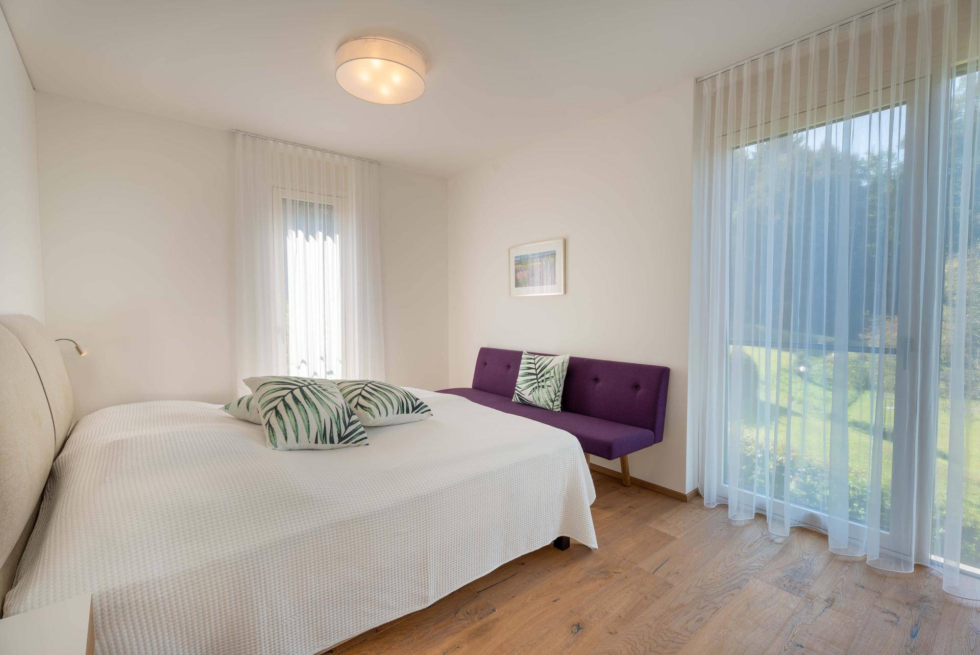 Helles Schlafzimmer mit großem Bett, lila Sofa und bodentiefen Fenstern mit Blick ins Grüne.