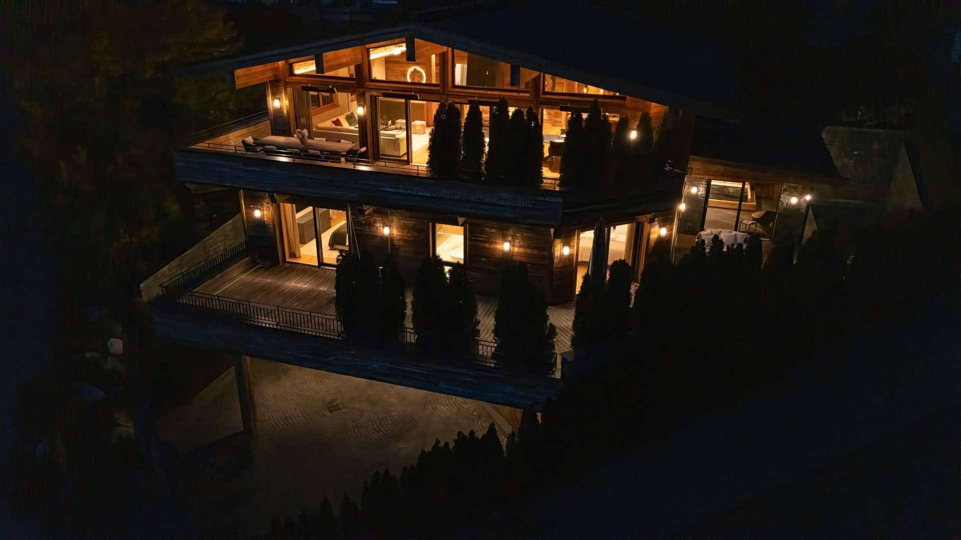 Luxuriöses Chalet bei Nacht, beleuchtet mit mehreren Terrassen und großen Glasfronten, eingebettet in die Natur.
