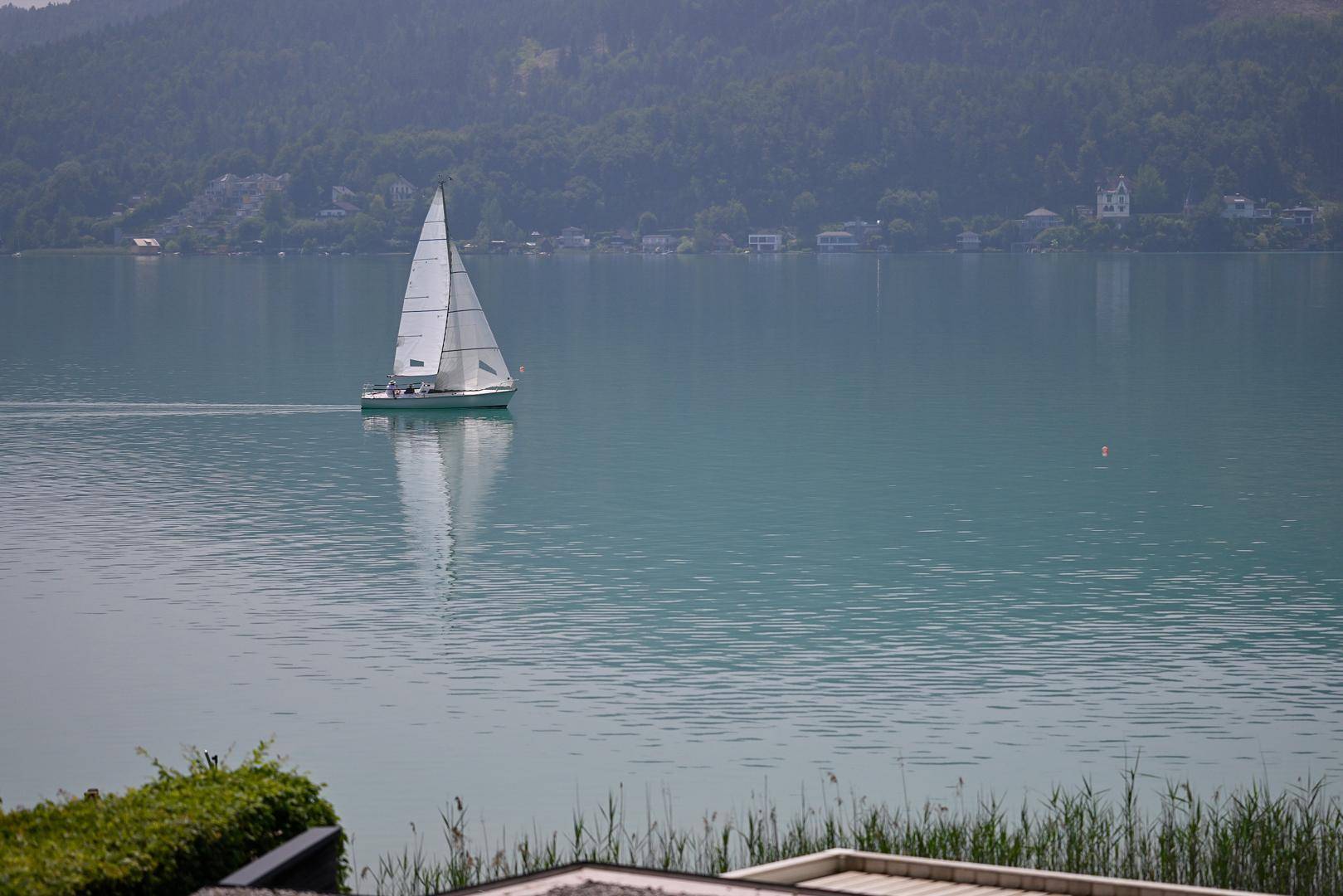 Segelboot auf dem ruhigen See mit bewaldeten Bergen und Häusern am Ufer im Hintergrund an einem sonnigen Tag.