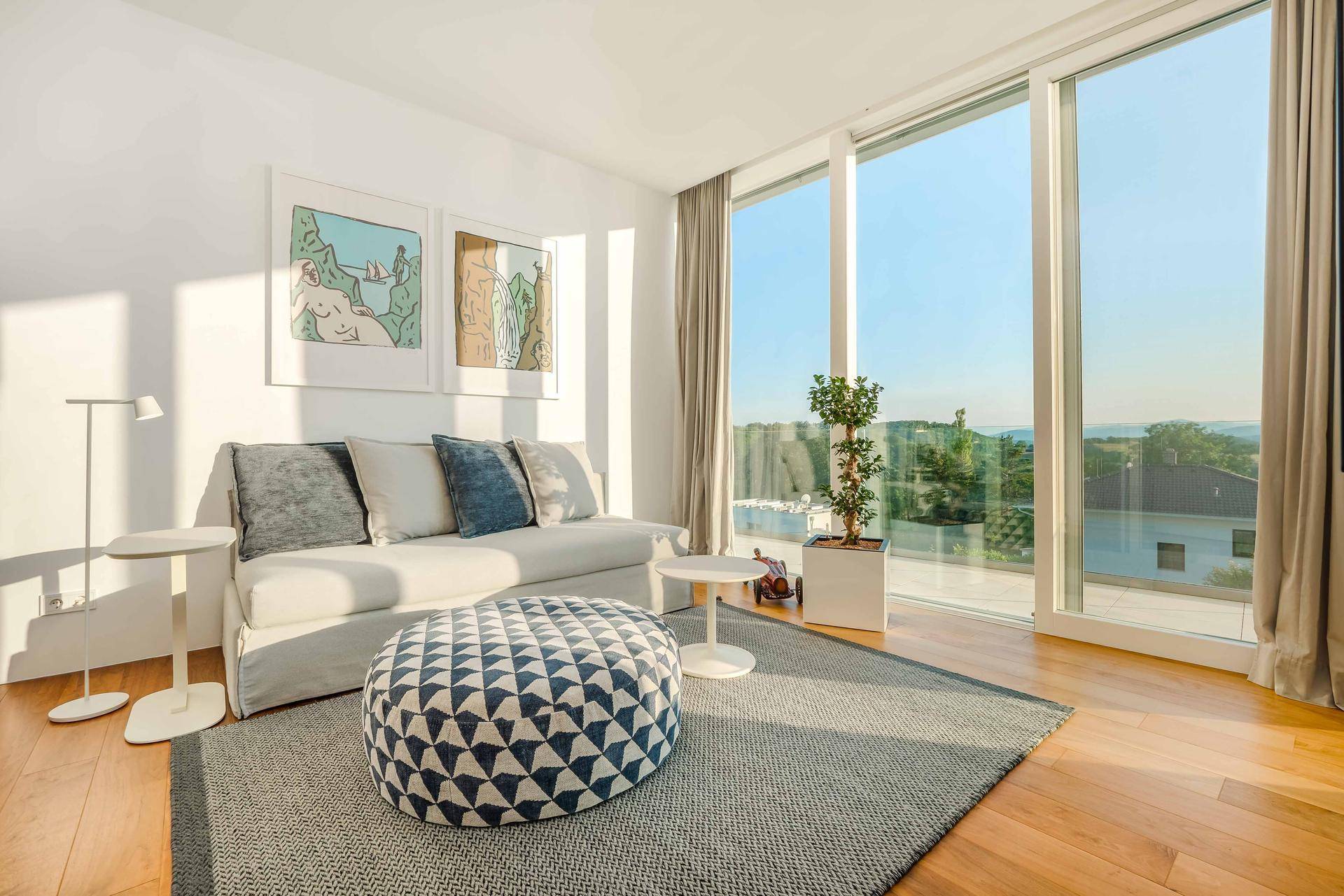 Lichtdurchflutetes Wohnzimmer mit weißem Sofa, Holzboden und großen Fenstern, die einen weiten Ausblick ermöglichen.