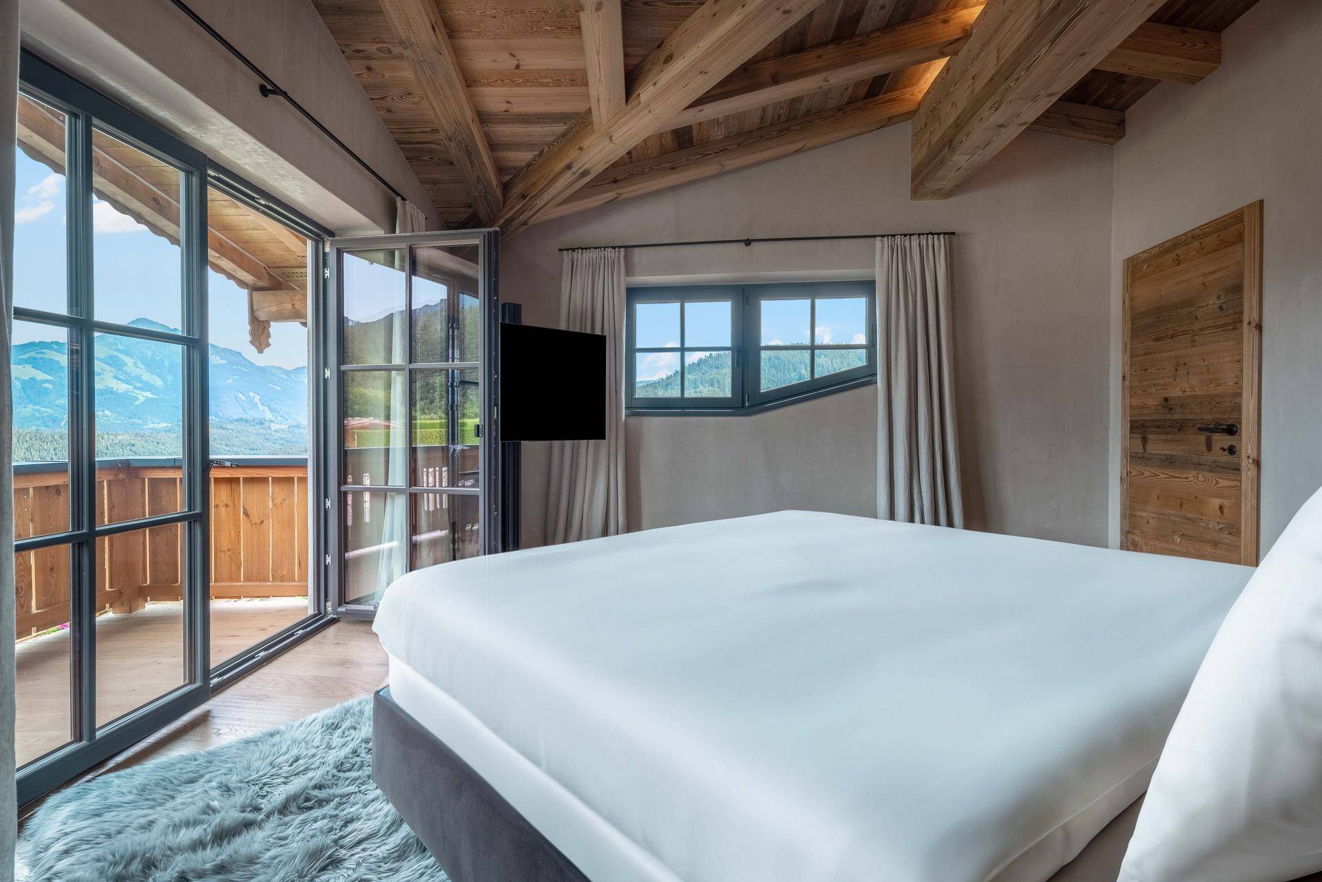Helles Schlafzimmer mit Holzbalken, großem Bett und Zugang zum Balkon mit Bergblick.
